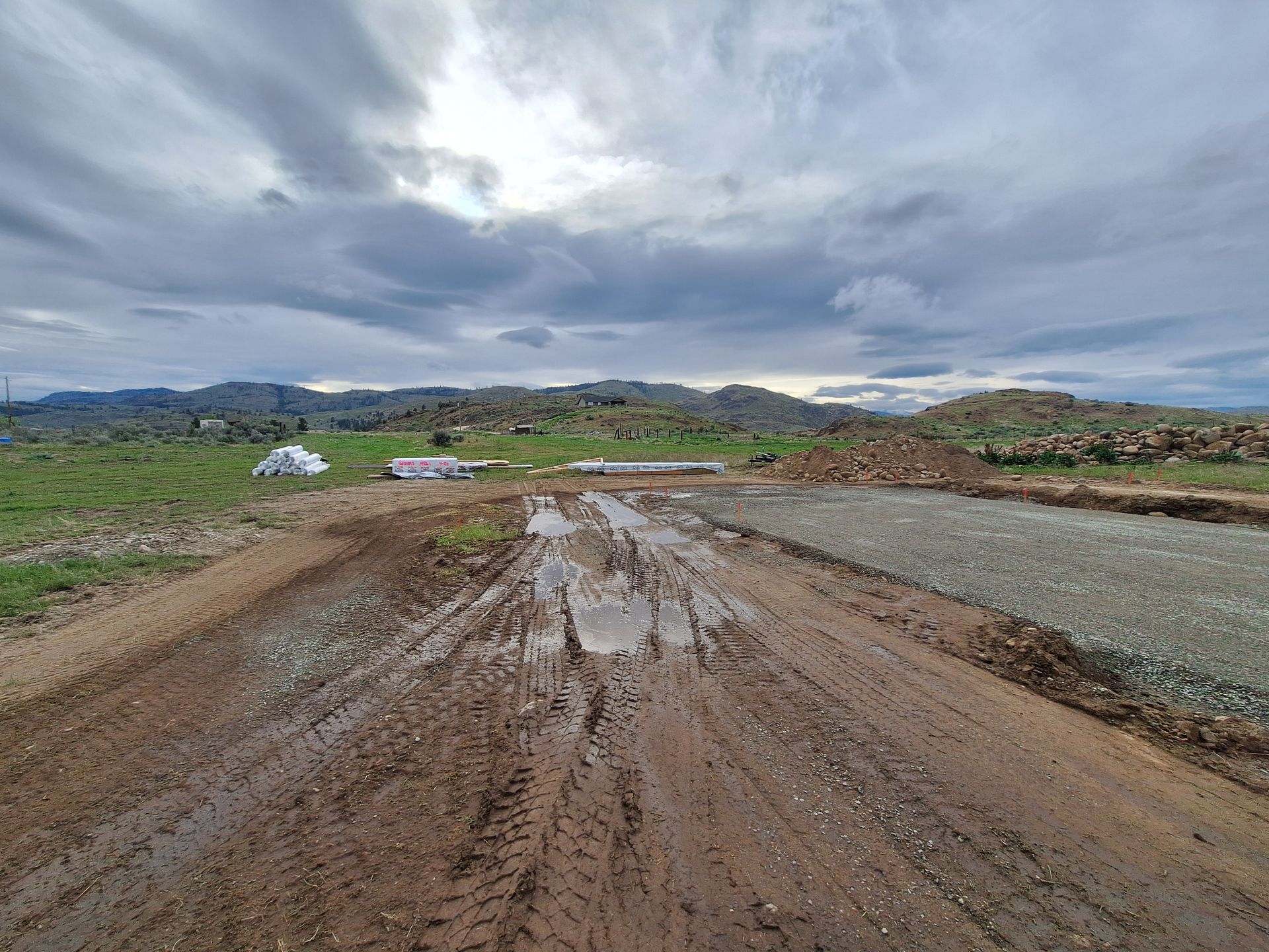Muddy dirt road leads to construction site under cloudy sky.