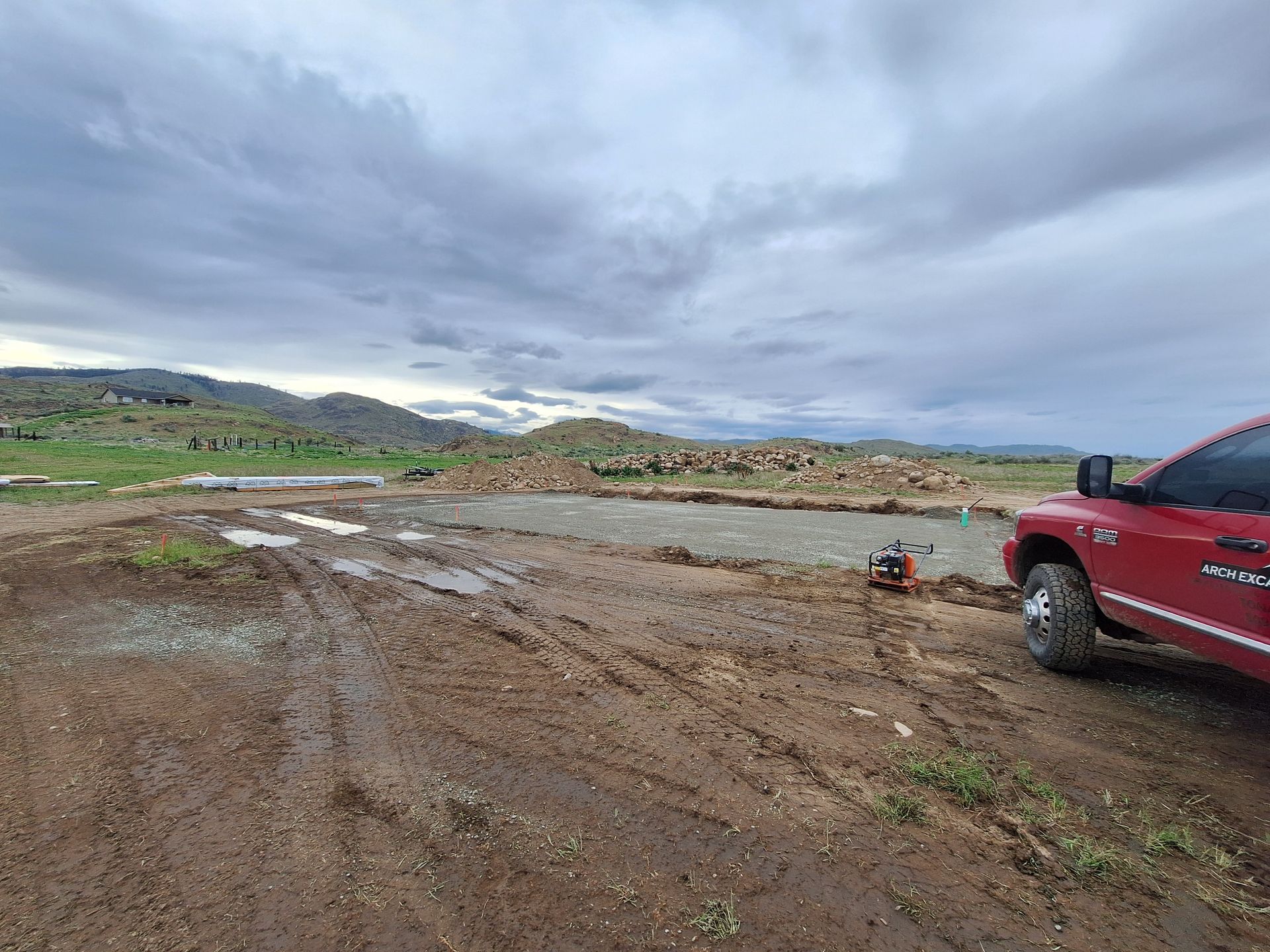 Muddy construction site; red truck parked on right, cloudy sky, dirt, and distant hills.
