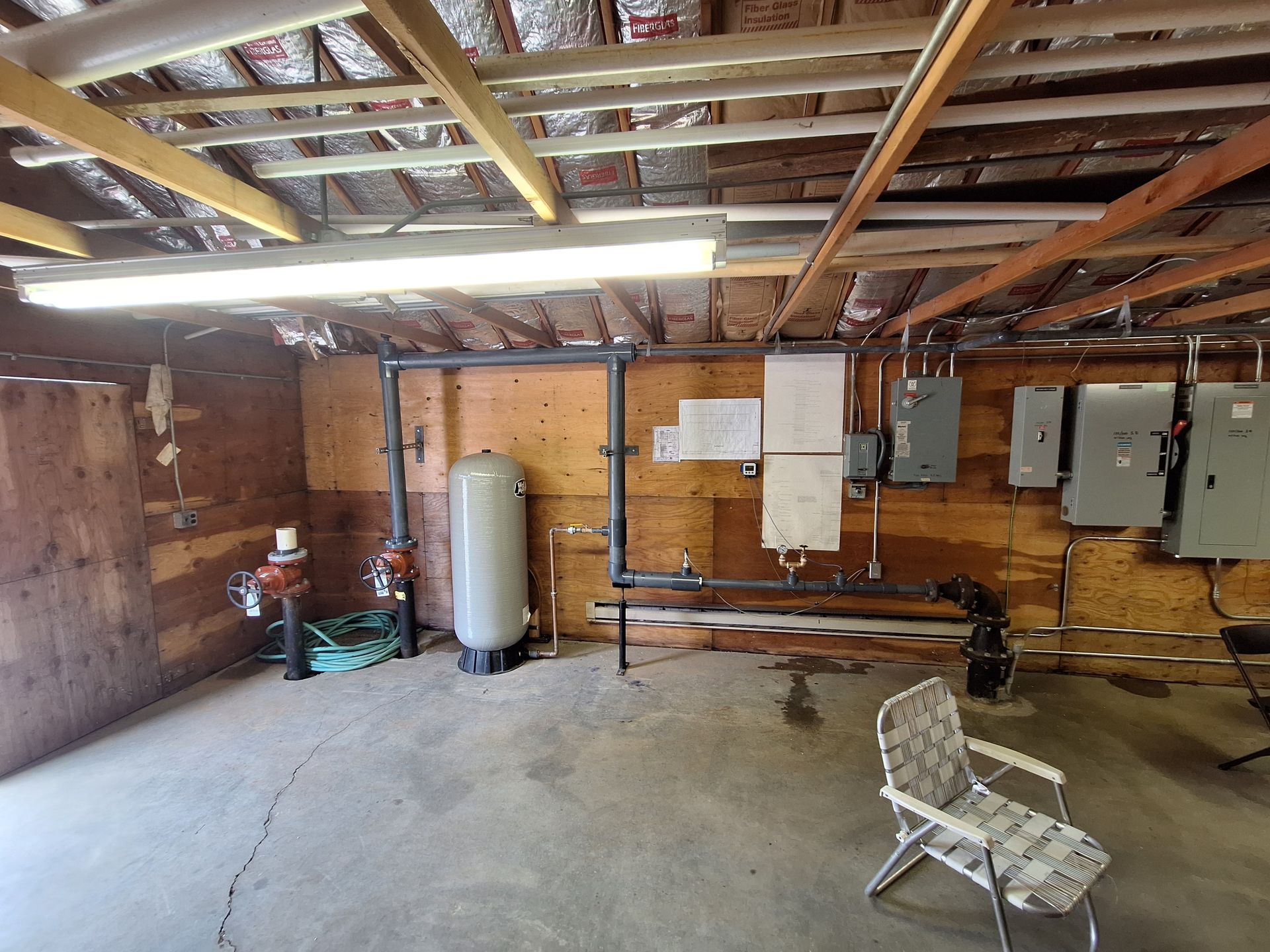 Interior of a utility room with plumbing, electrical panels, and a water tank. Concrete floor, wooden walls, exposed ceiling.