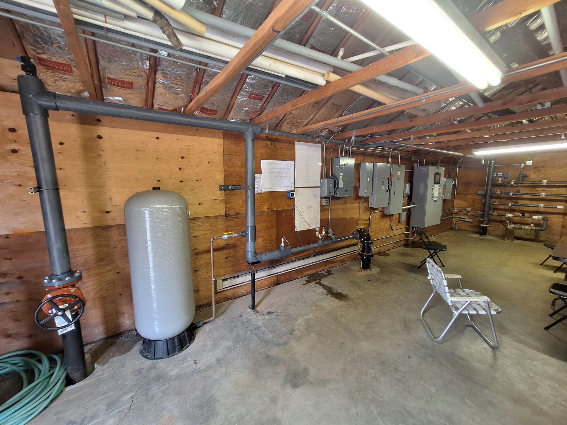A water system room with pipes, tank, and electrical panels. Wooden walls and concrete floor.