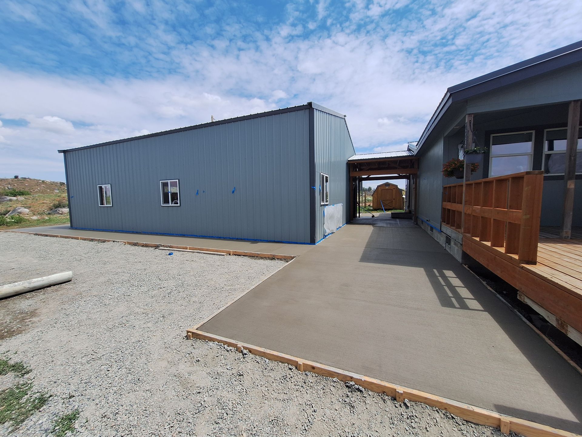 Blue metal building connected by a covered walkway to a house with a wooden deck; gravel and concrete path.