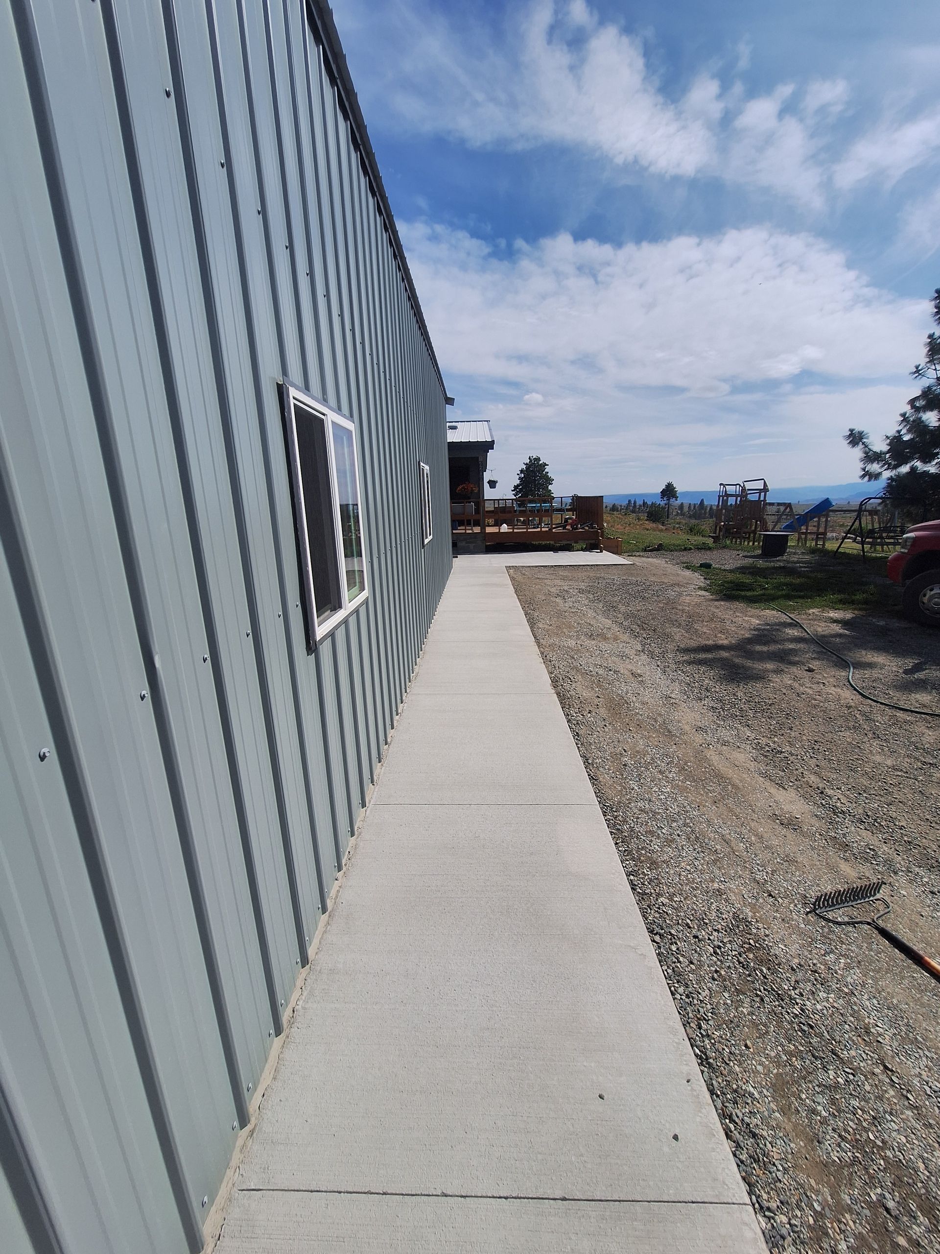 Concrete walkway alongside a gray metal-sided building, leading towards a wooden deck and distant landscape under a partly cloudy sky.