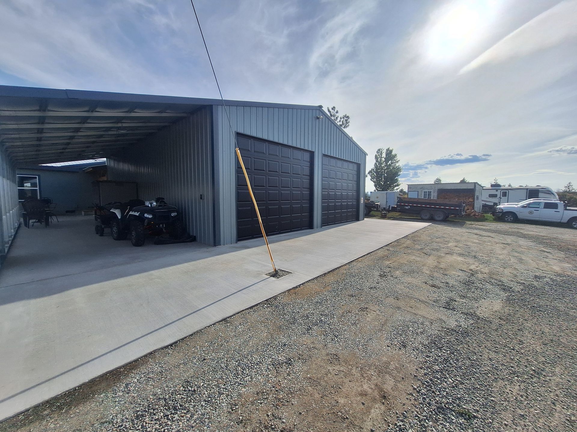Metal garage with black doors and concrete pad; motorcycles parked inside. Gravel and parked vehicles visible.