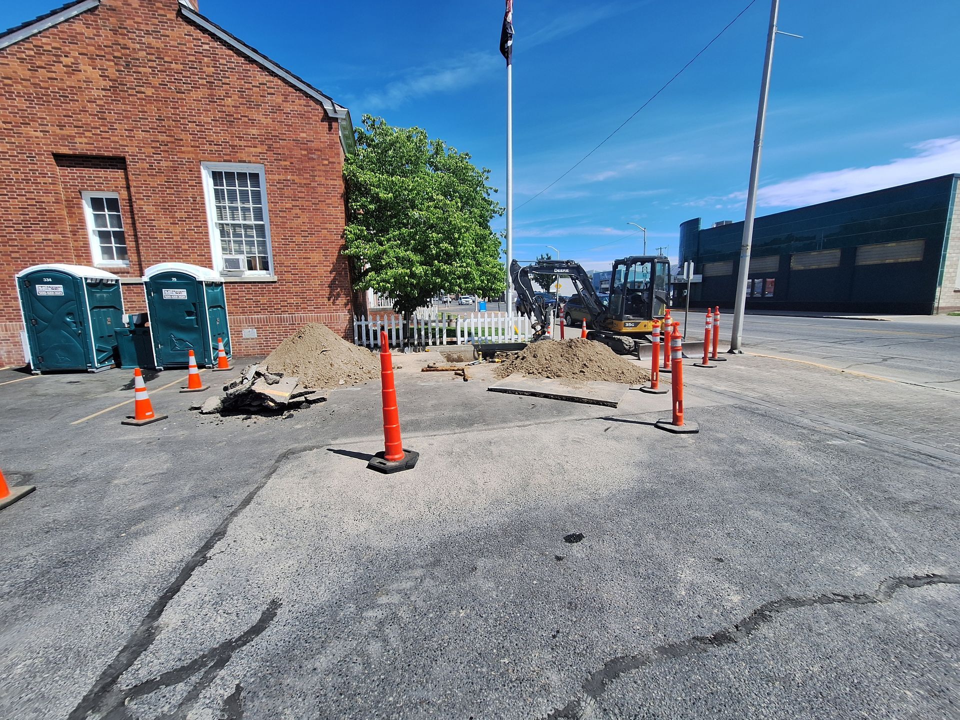 Construction site on paved area next to brick building. Backhoe, dirt piles, and orange safety cones are present.