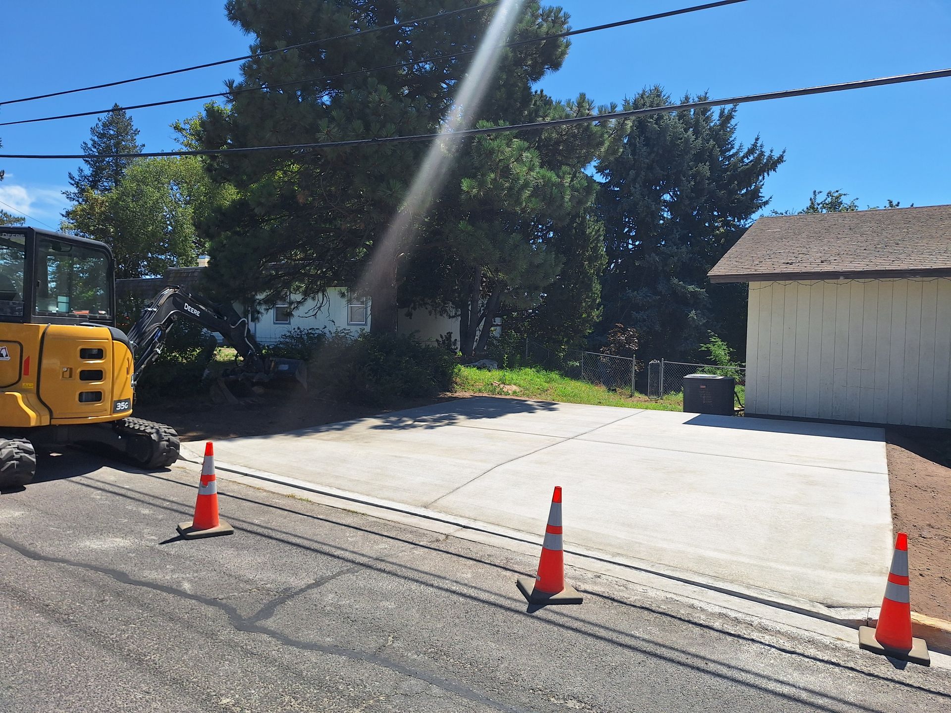Yellow excavator on a paved area with orange cones, driveway, small building, trees, and power lines.