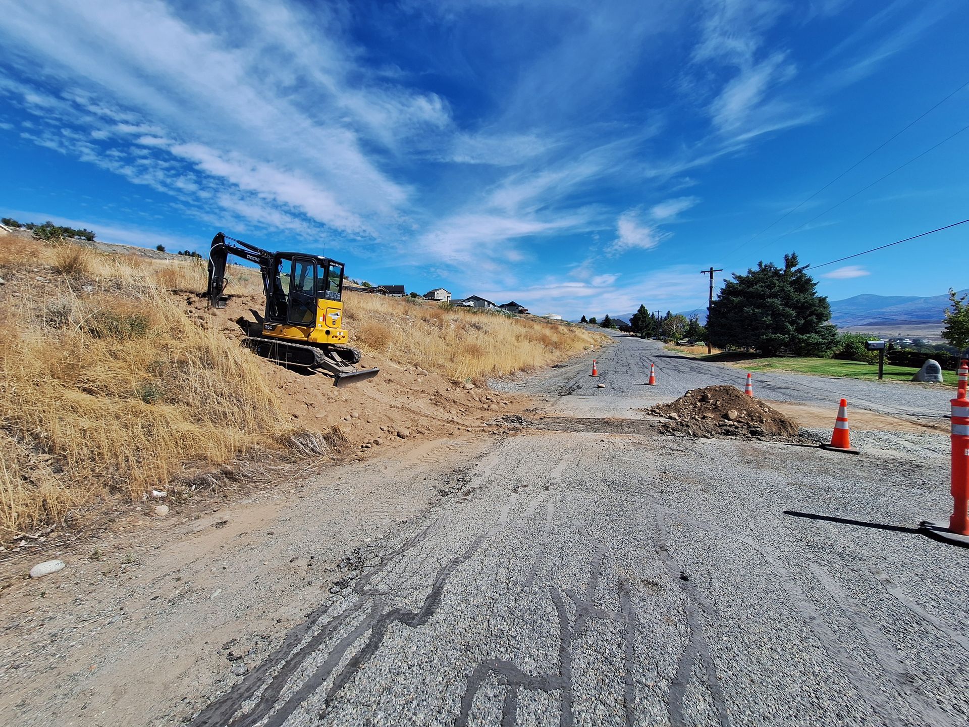 Excavator on a hillside clearing dirt. Gravel path in foreground, orange traffic cones. Blue sky.