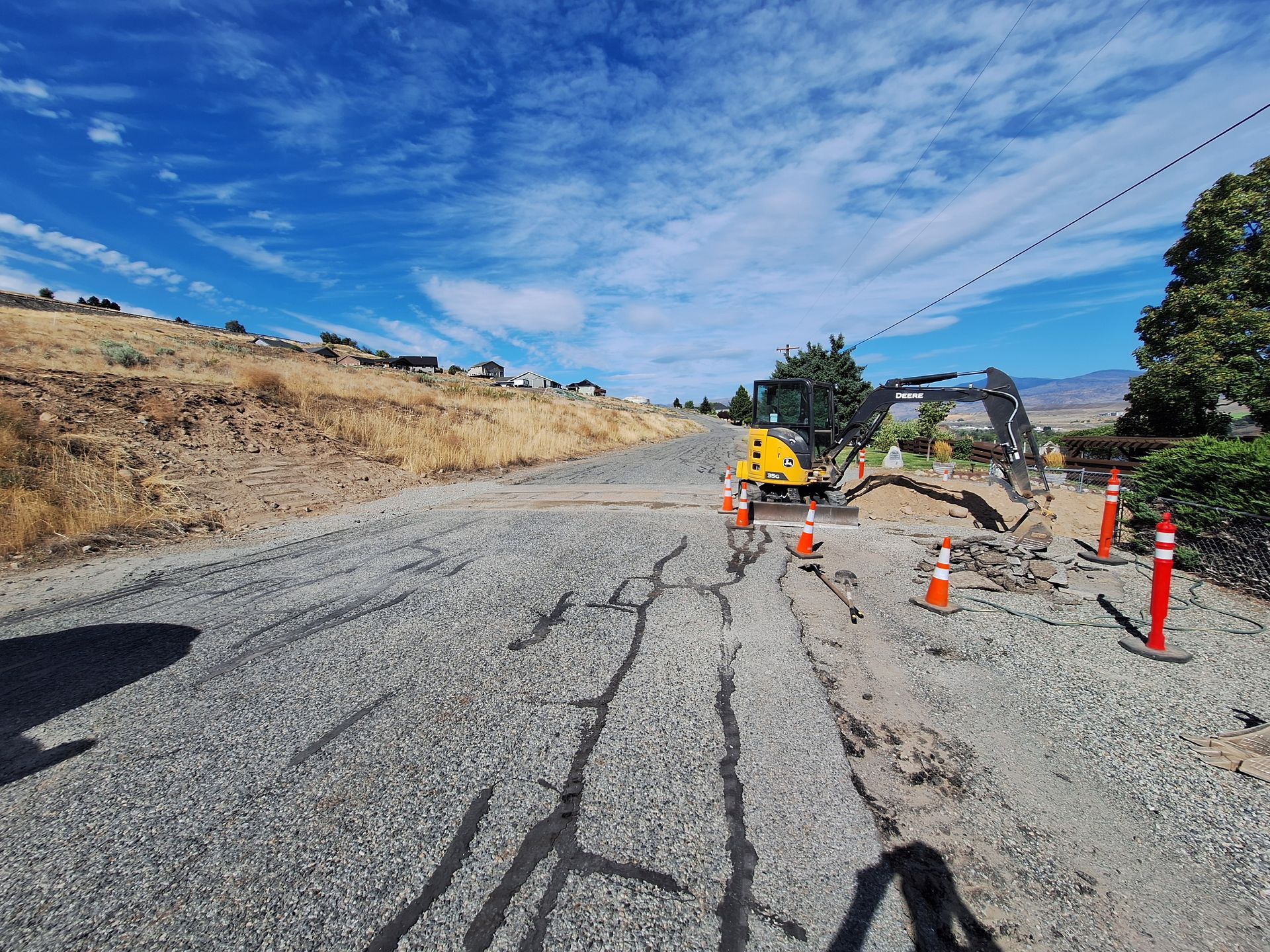 A small excavator working on a gravel road, orange cones marking the area, under a blue sky.