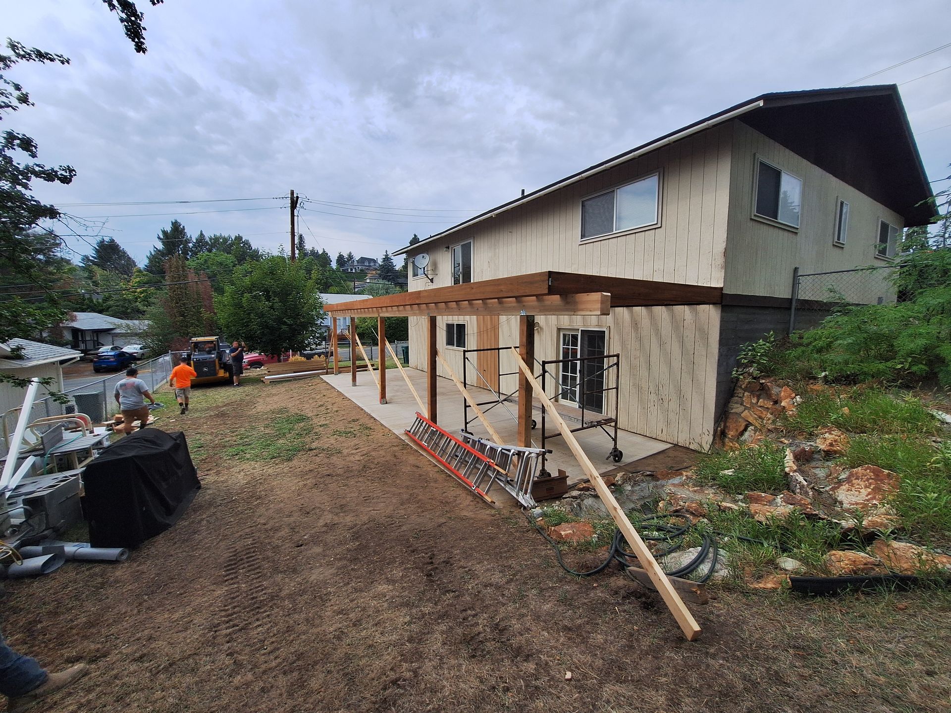 Construction of a wooden pergola attached to a two-story beige house. Workers and equipment visible. Overcast day.