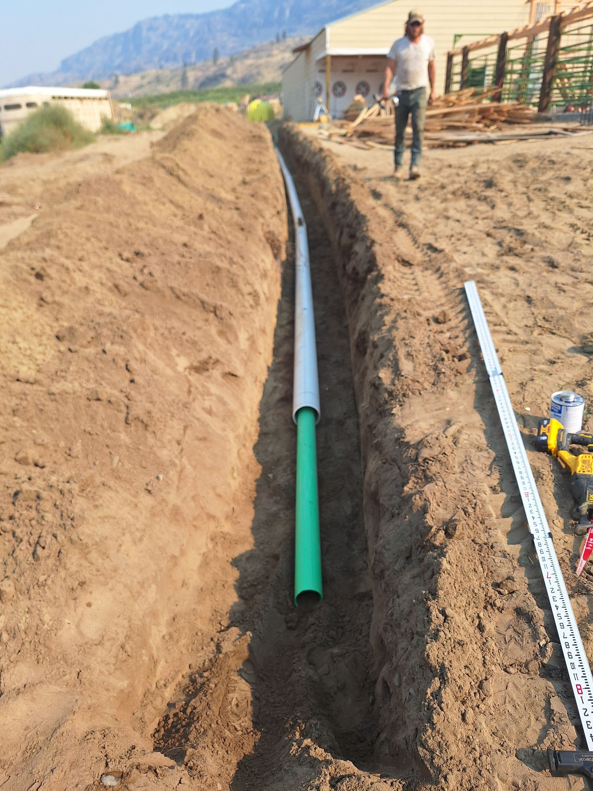 Trench with green and white pipes, a worker, and a building on a dirt lot.