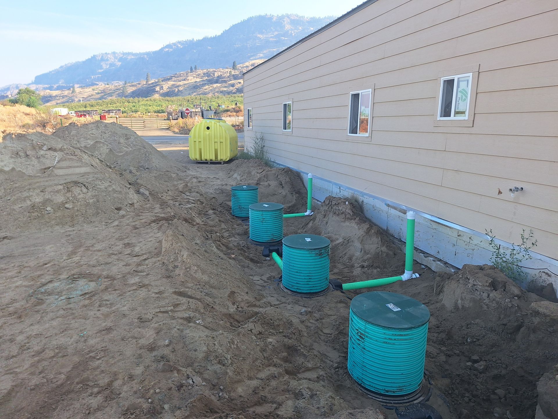 Septic system installation alongside a beige building; green tanks, pipes, and yellow cover in dirt.