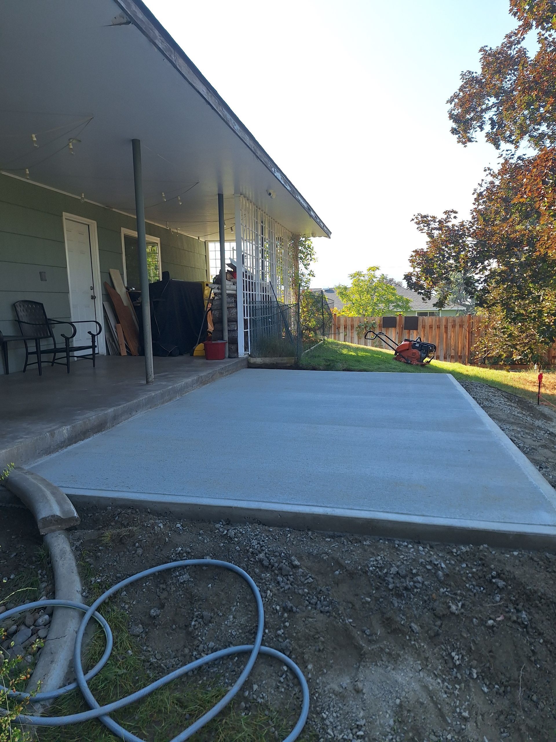 Newly poured concrete patio next to a house with an existing covered porch. Gravel border, hose visible.