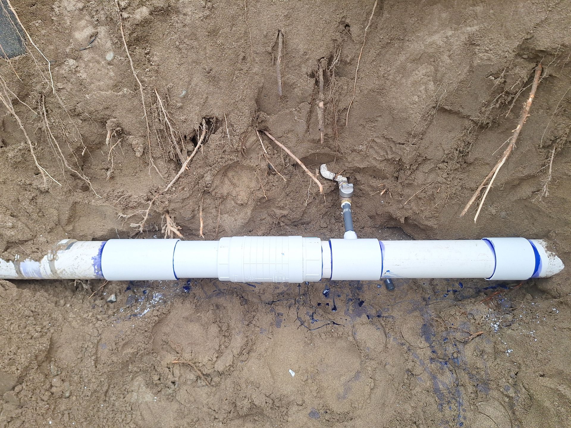 White PVC pipe sections with fittings partially buried in brown soil. A small valve is connected.