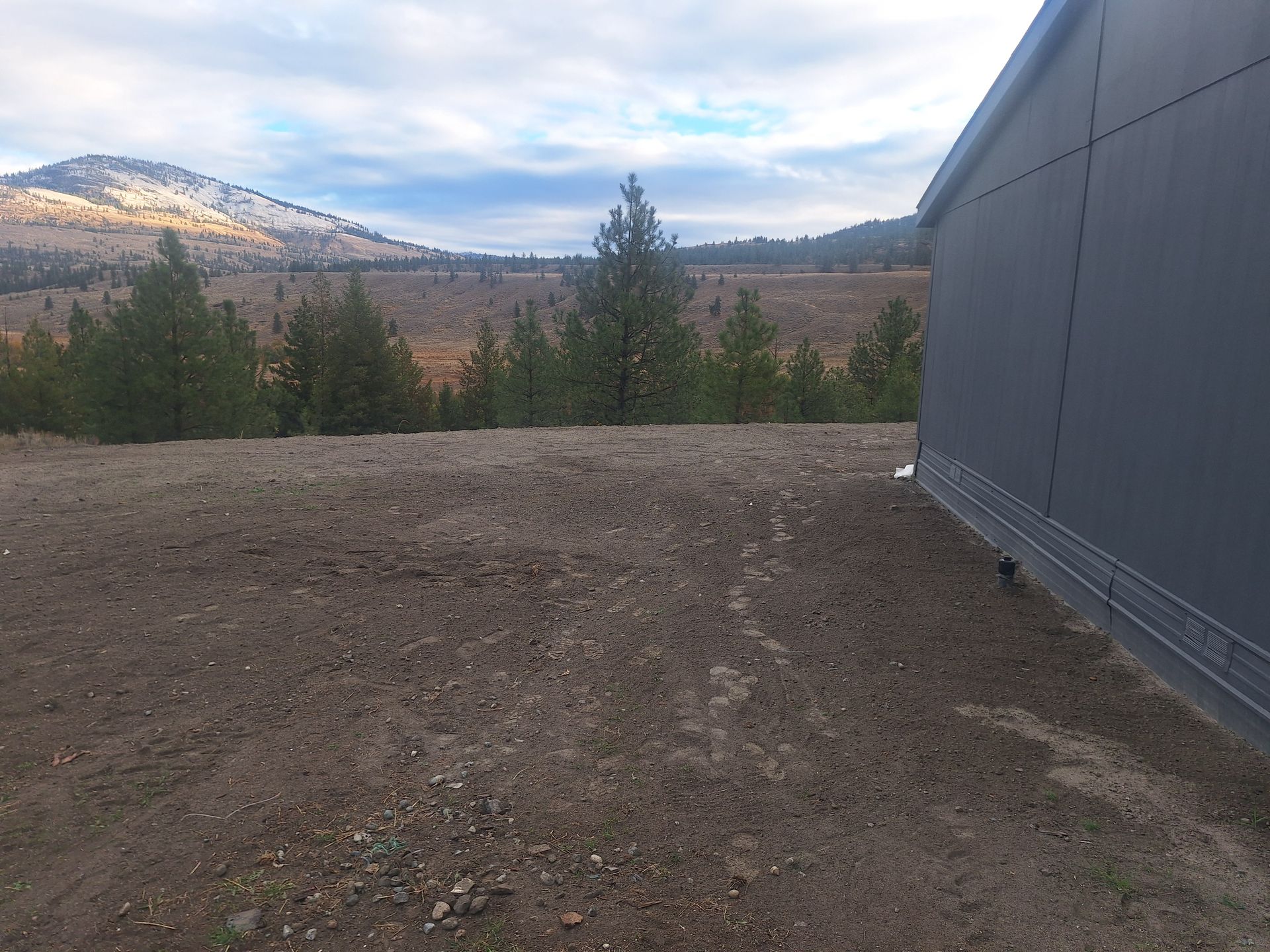 Dirt field and trees with a mountain backdrop, a gray building to the right.