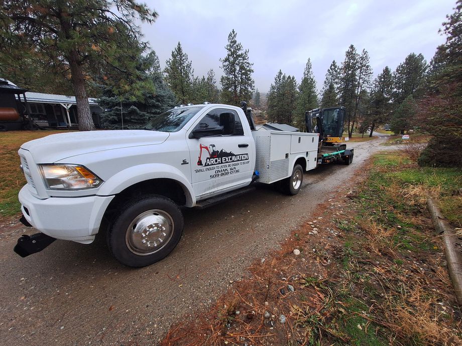 White work truck with a small excavator on a trailer on a gravel road, overcast day.