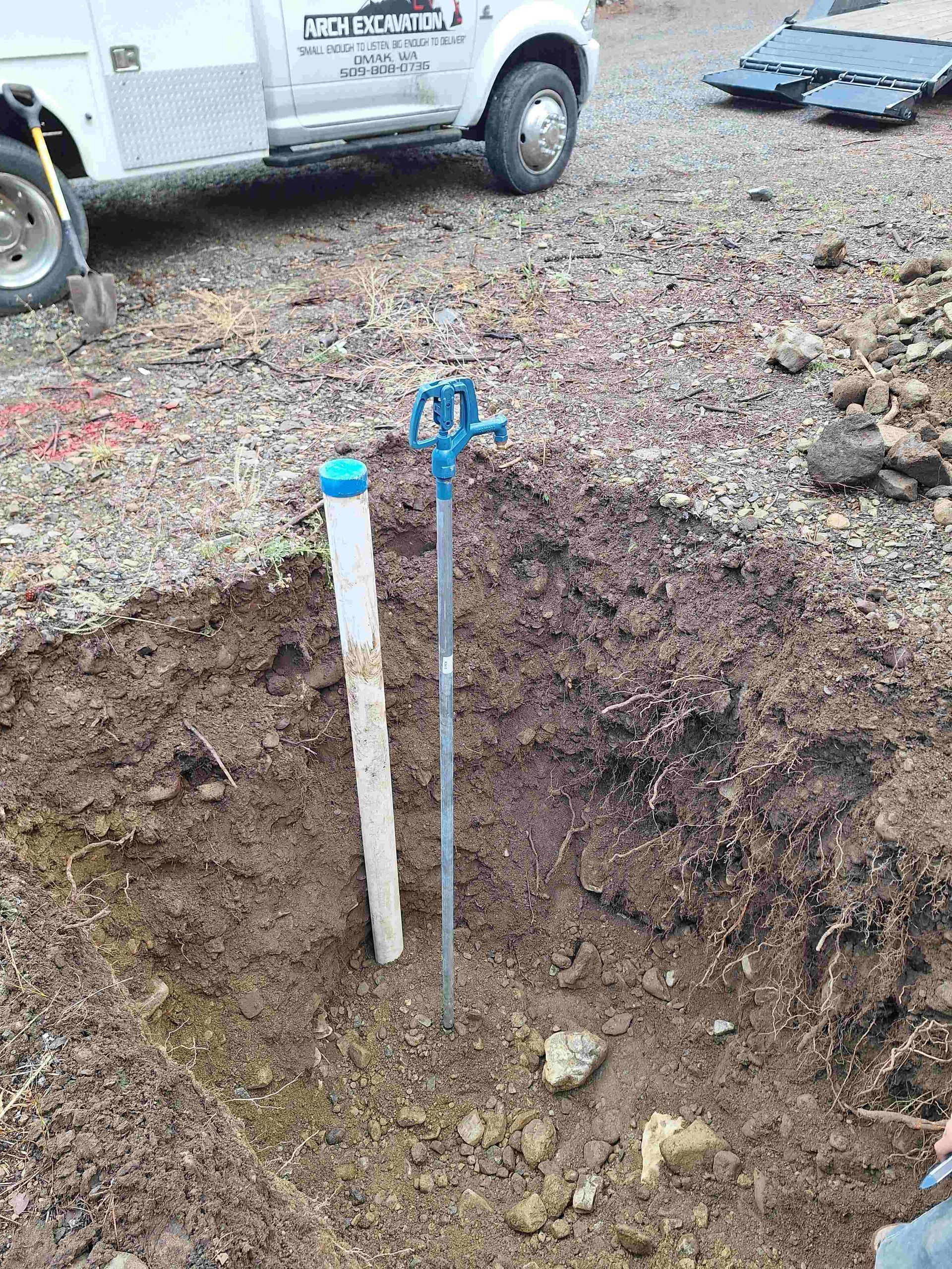 Excavated hole with white pipe and blue-topped hydrant in the ground, truck in background.