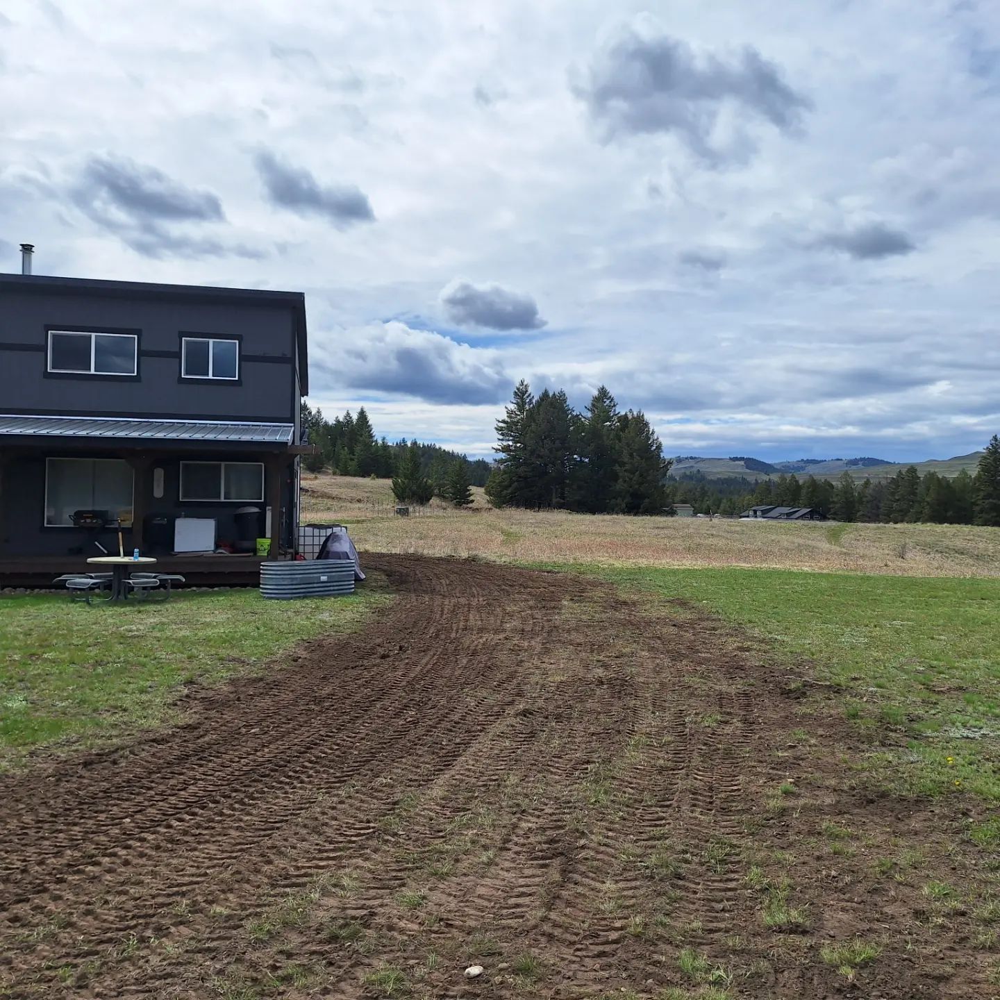 A dark house next to a dirt path in a grassy field with trees and mountains in the distance under a cloudy sky.
