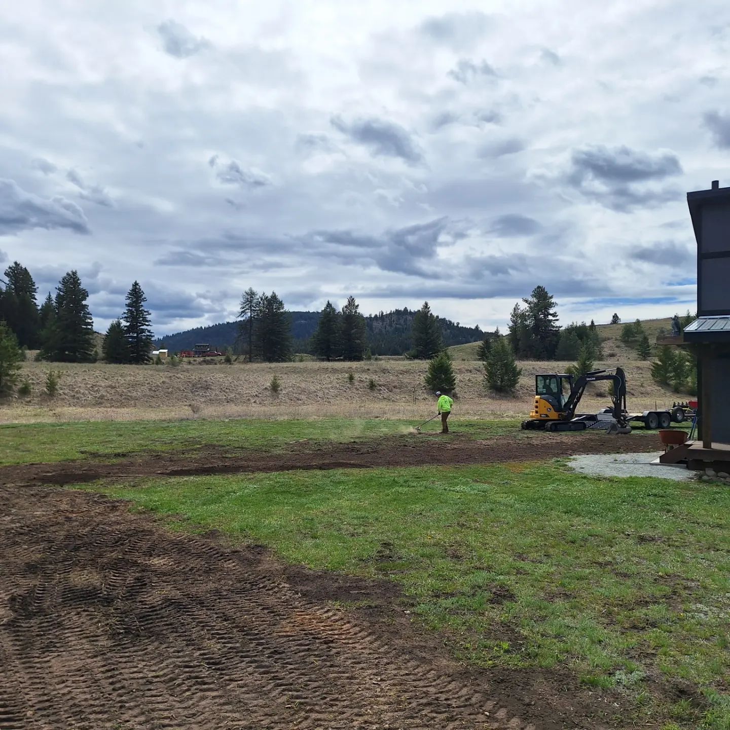 Construction site with excavator and worker in a field; trees and cloudy sky in the background.