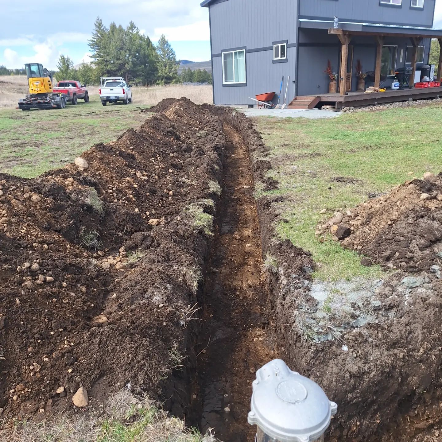 A trench dug in a grassy yard, leading to a utility box near a house under construction.