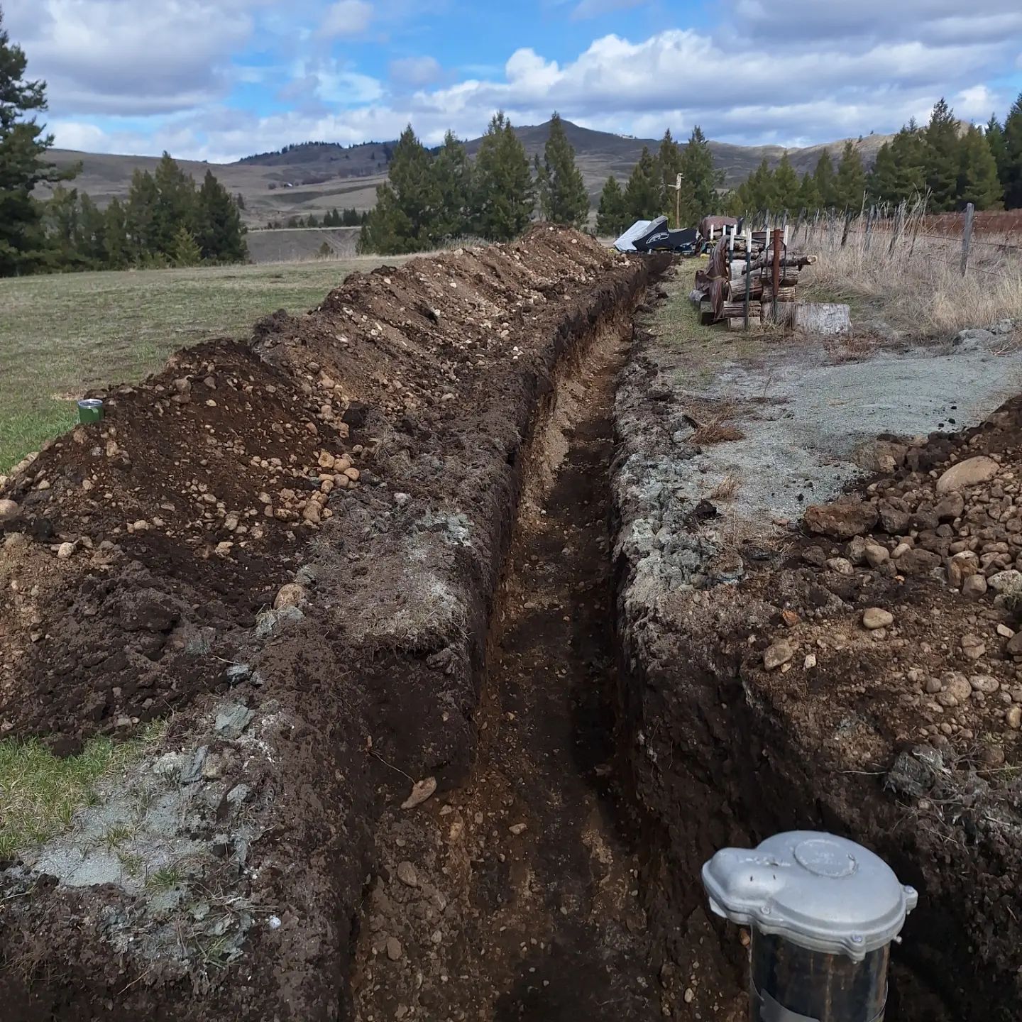 Long, narrow dirt trench in a field, leading towards pipes and a hillside. Cloudy sky.