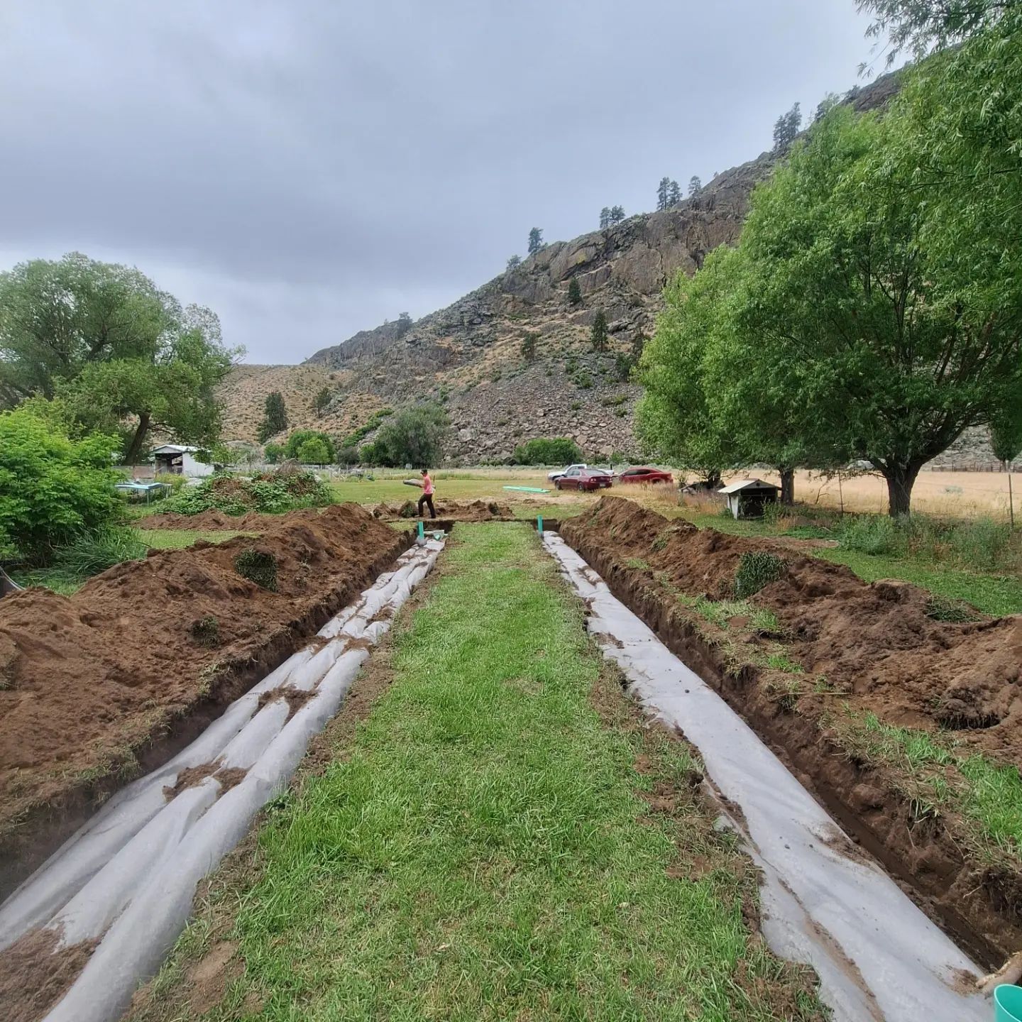 Trenches dug in a grassy field, lined with white fabric. A person works in the distance, mountain backdrop.