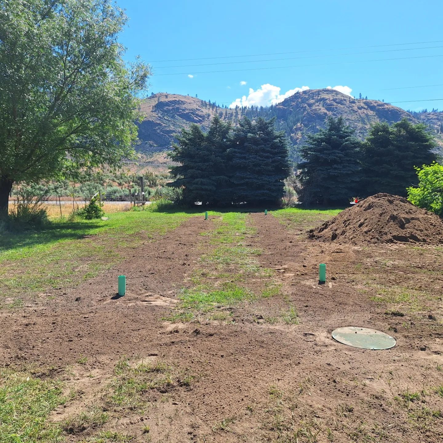 Cleared yard with green markers, septic tank, and dirt mound, trees, and mountains in background.
