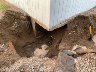 Excavation around a building's corner. Dark soil and gravel surround a white-sided structure. A yellow shovel rests nearby.