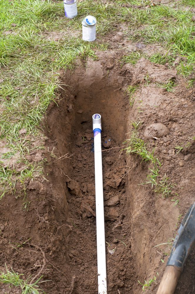 Trench dug in soil with white PVC pipe, two cans of primer/cement, and a shovel, outdoors.