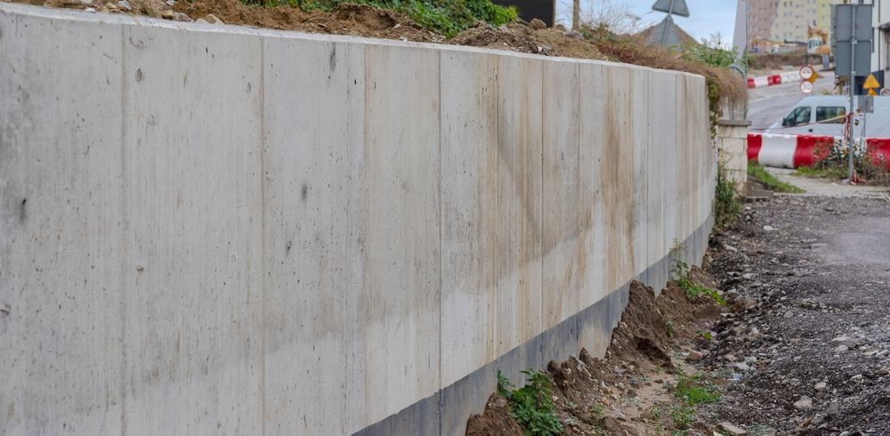 A concrete retaining wall along a road, with some dirt and grass at its base.