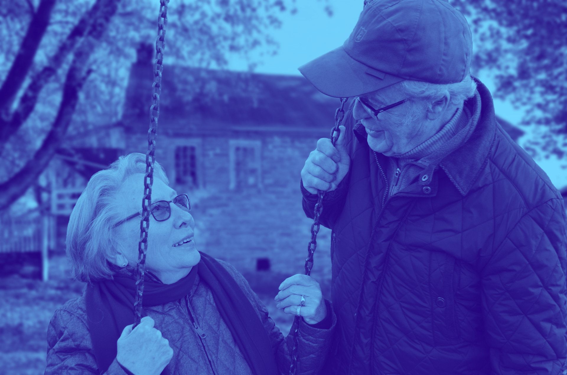 An elderly couple is sitting on a swing in a park.