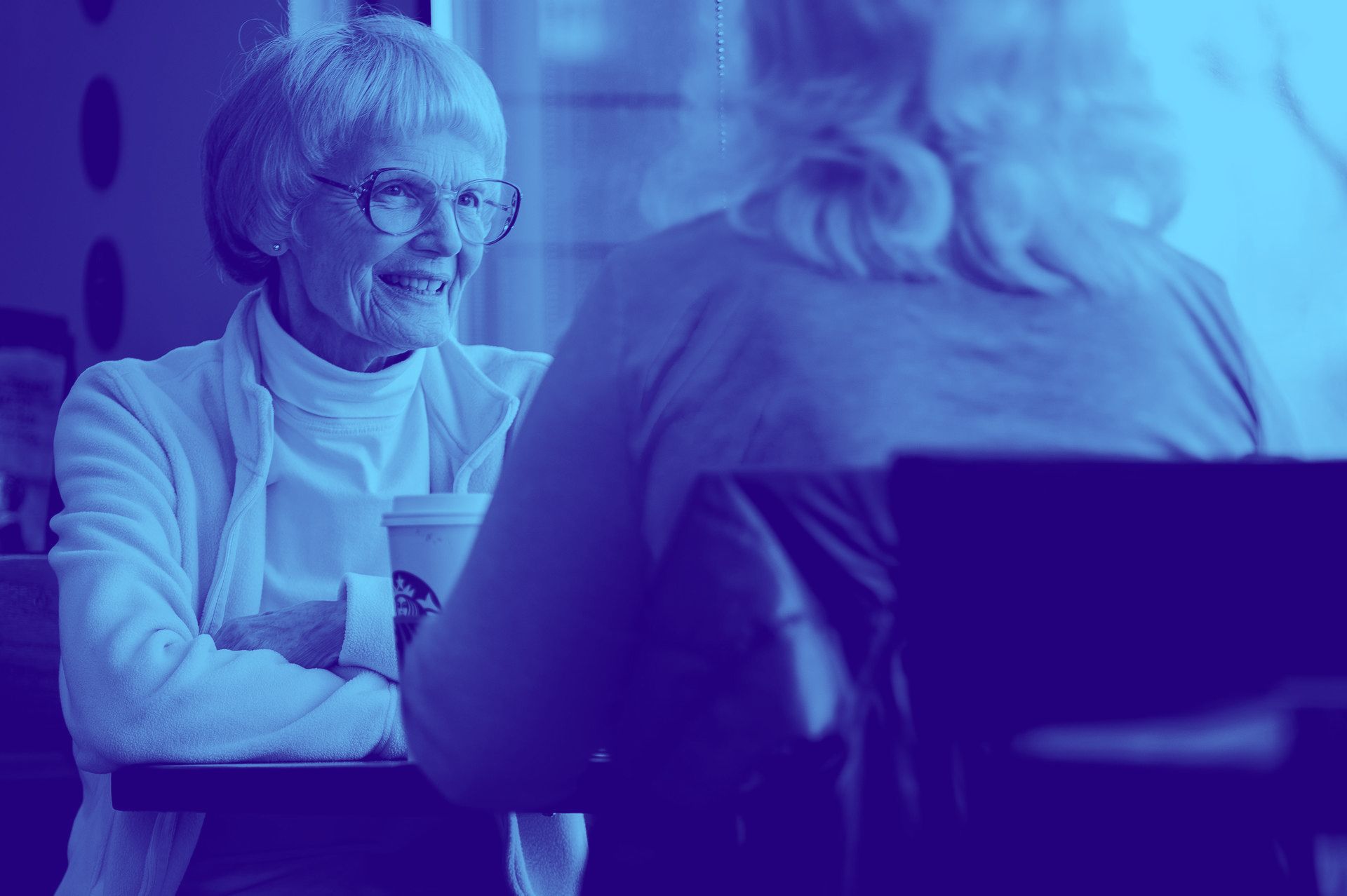 Two older women are sitting at a table talking to each other.