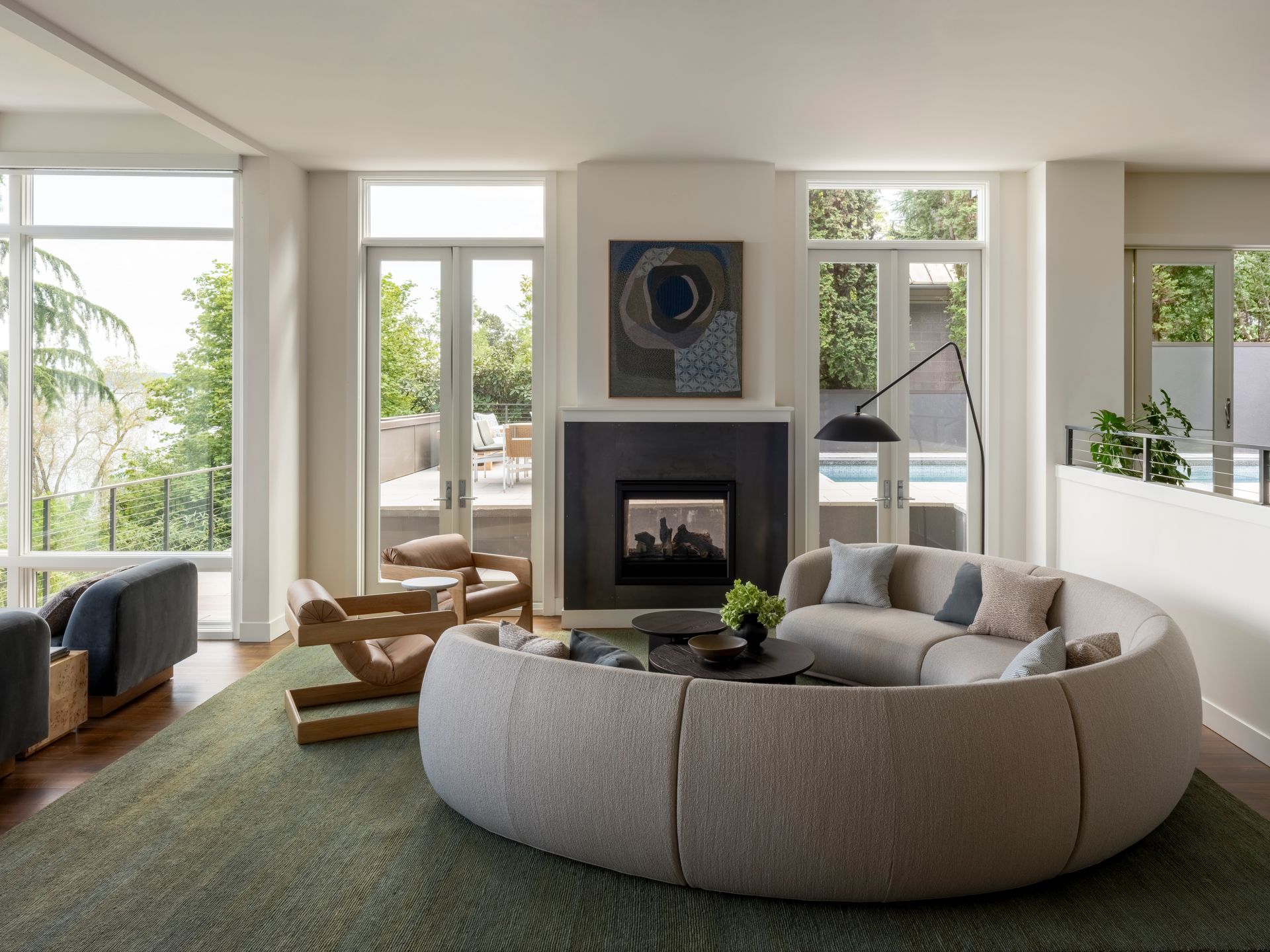 Living room with circular sofa, fireplace, and large windows overlooking greenery.