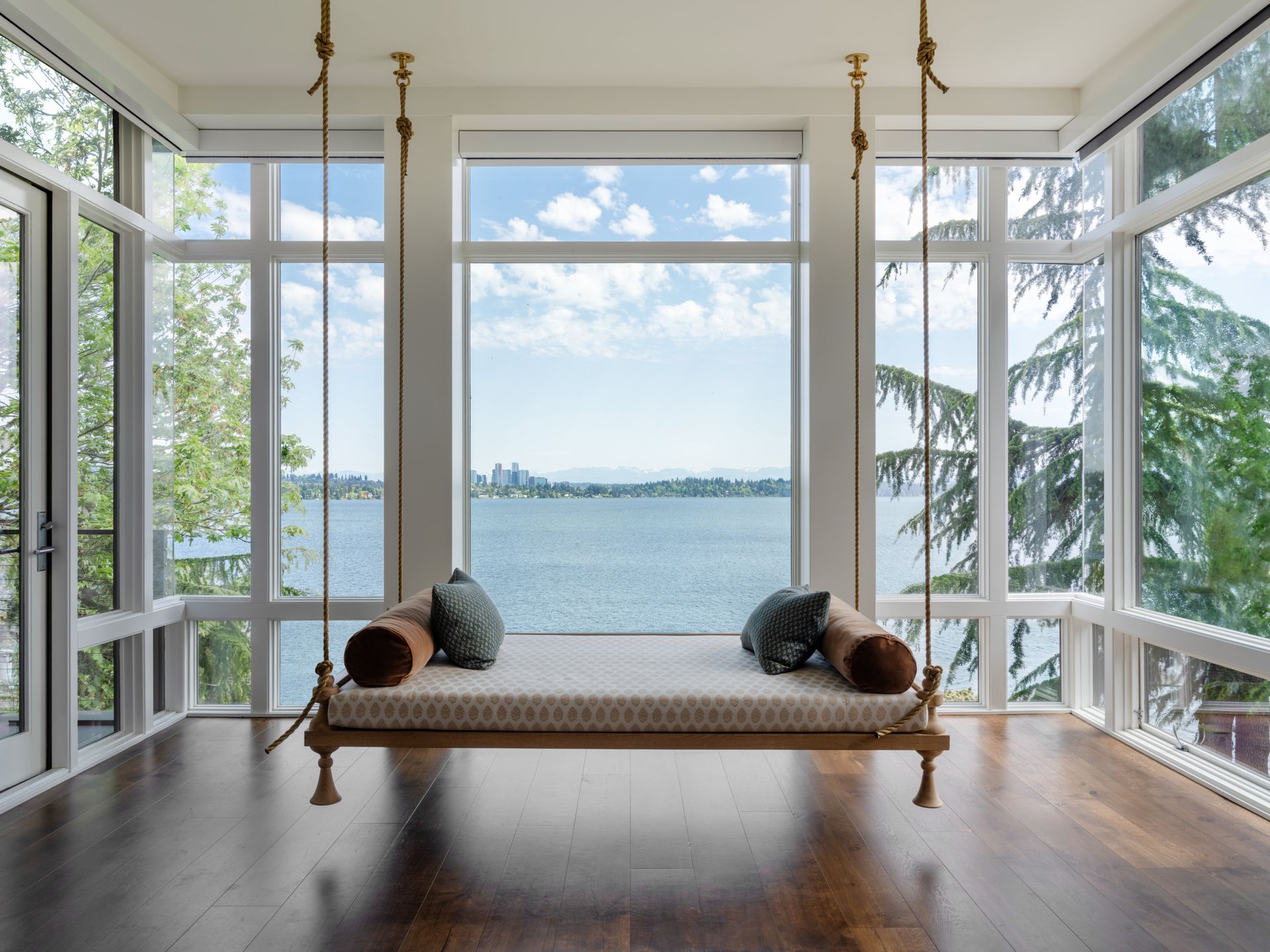 Wooden swing with patterned cushions, overlooking a scenic lake view with large windows.