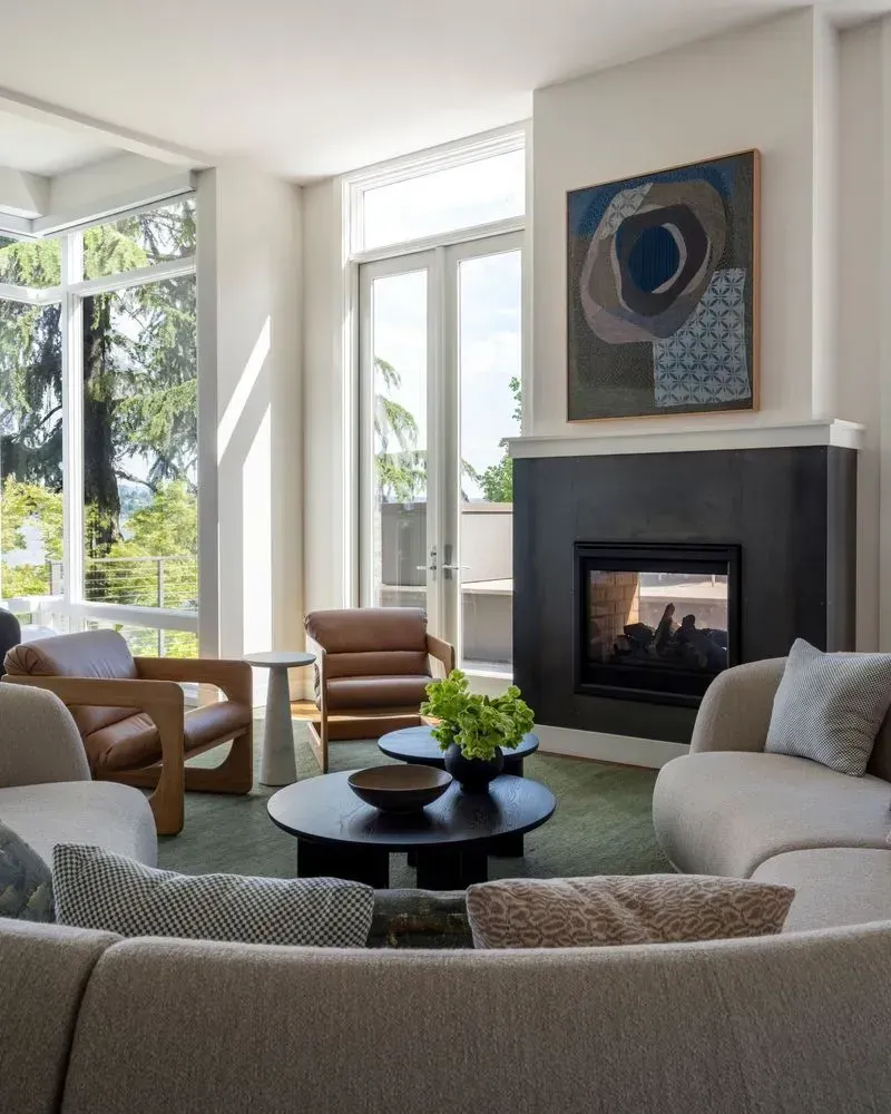 Living room with fireplace, round couch, brown leather chairs, round coffee table, artwork, and large windows.