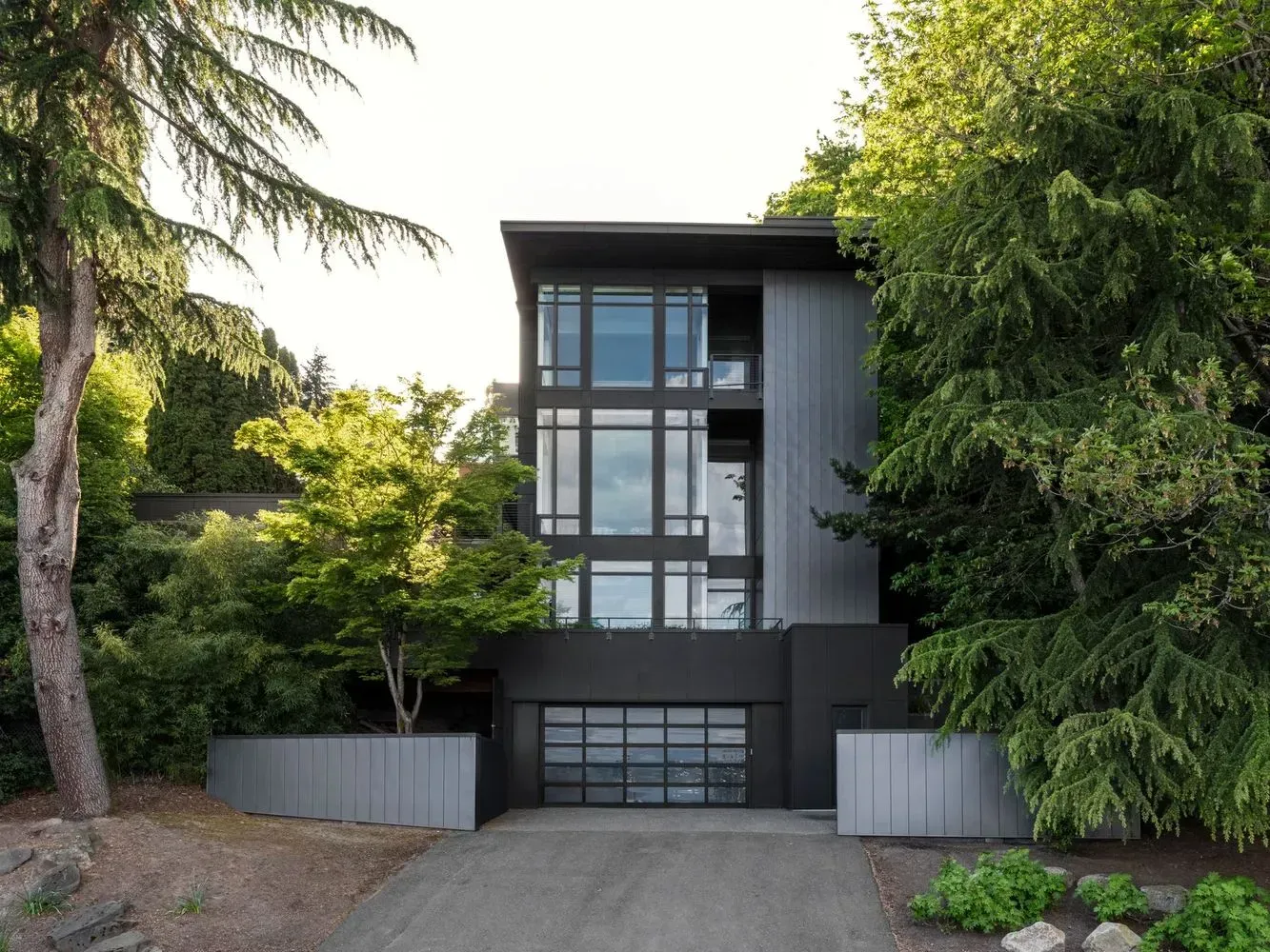 Modern black house with large windows, surrounded by green trees, a driveway, and gray fences.