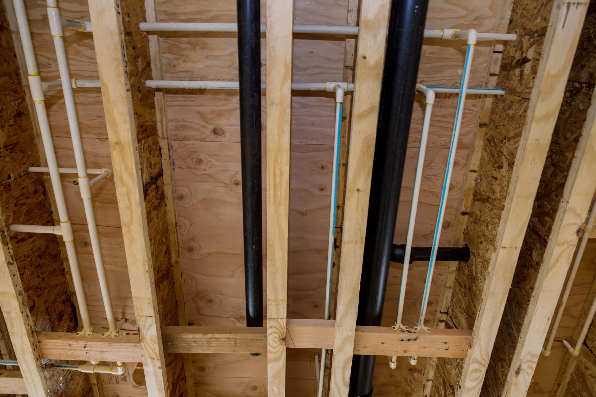 Pipes of various colors (white, black, blue) run through wooden ceiling joists in a construction setting.
