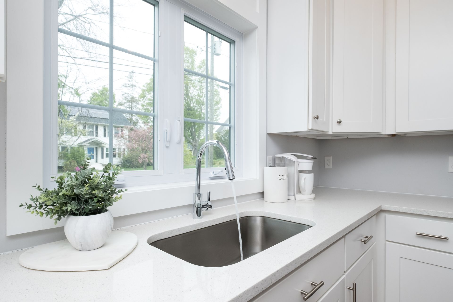White kitchen with sink under a window, green plant, and white cabinets; daylight.