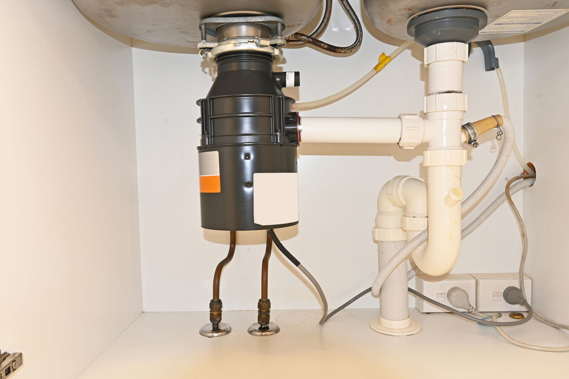 Under-sink view: garbage disposal, plumbing pipes, and electrical outlet in a white cabinet.