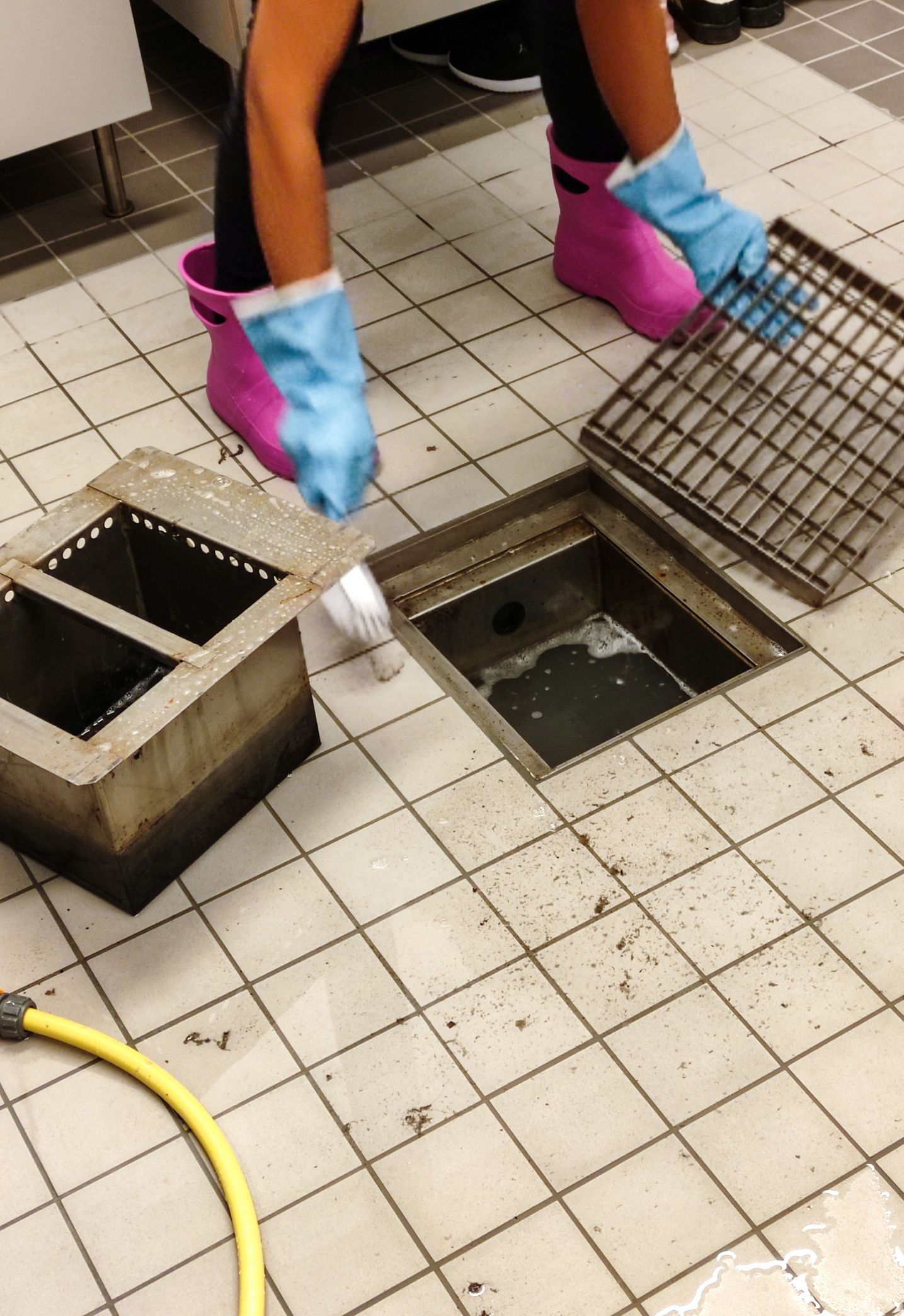 Person cleaning a floor drain, wearing pink and blue protective boots. Kitchen setting with a yellow hose.