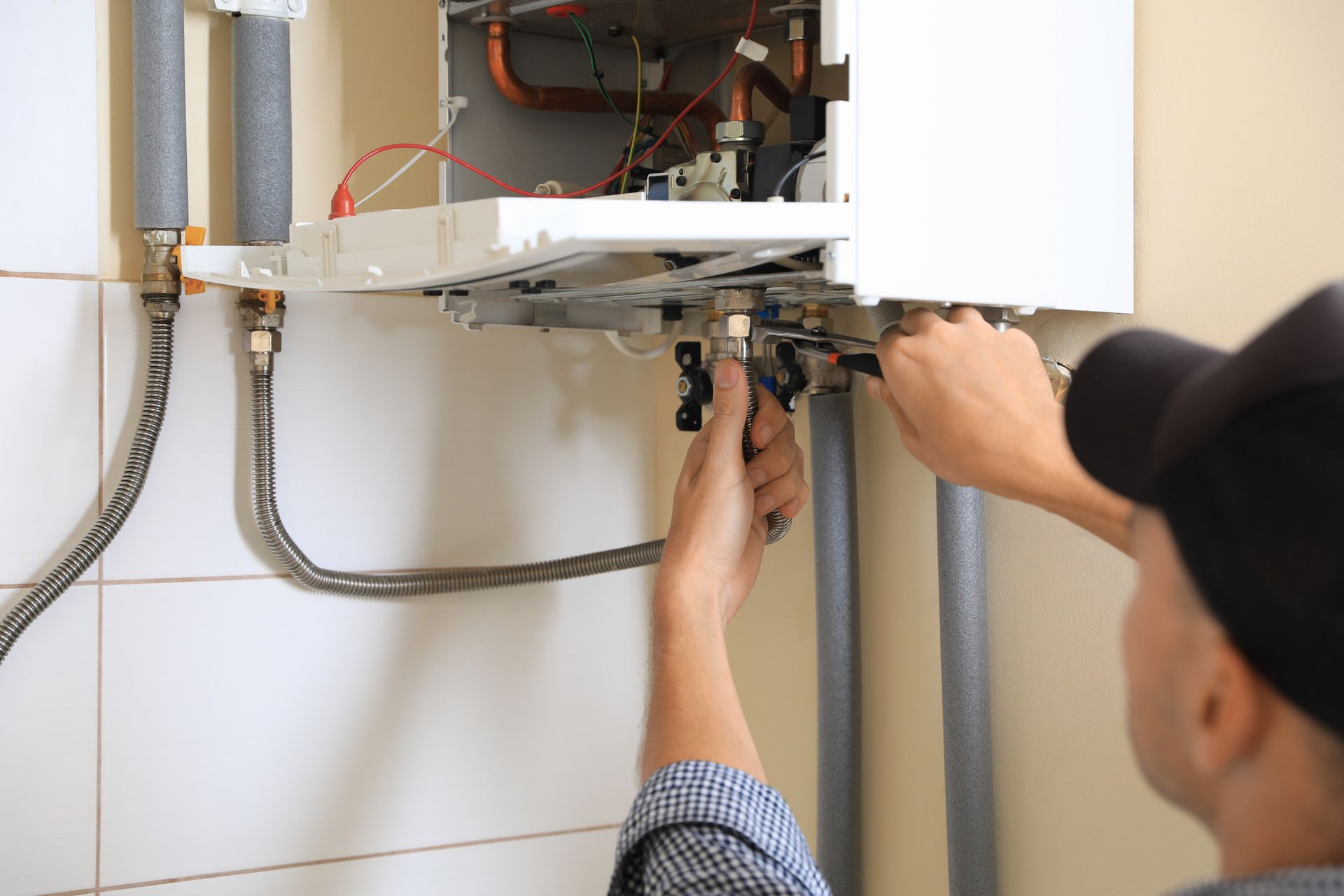A person in a black cap repairs a white boiler with a wrench. Indoor setting.
