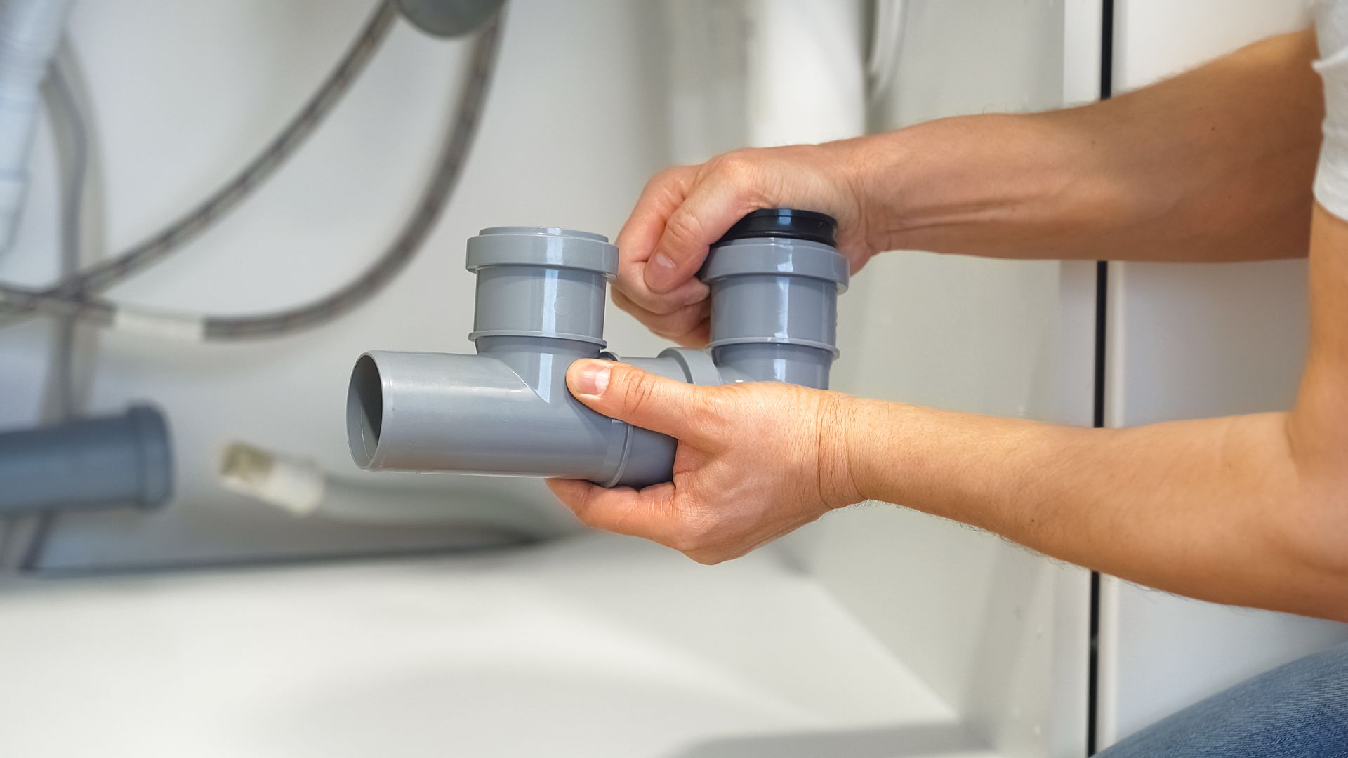 Hands assembling gray PVC pipes under a white sink.
