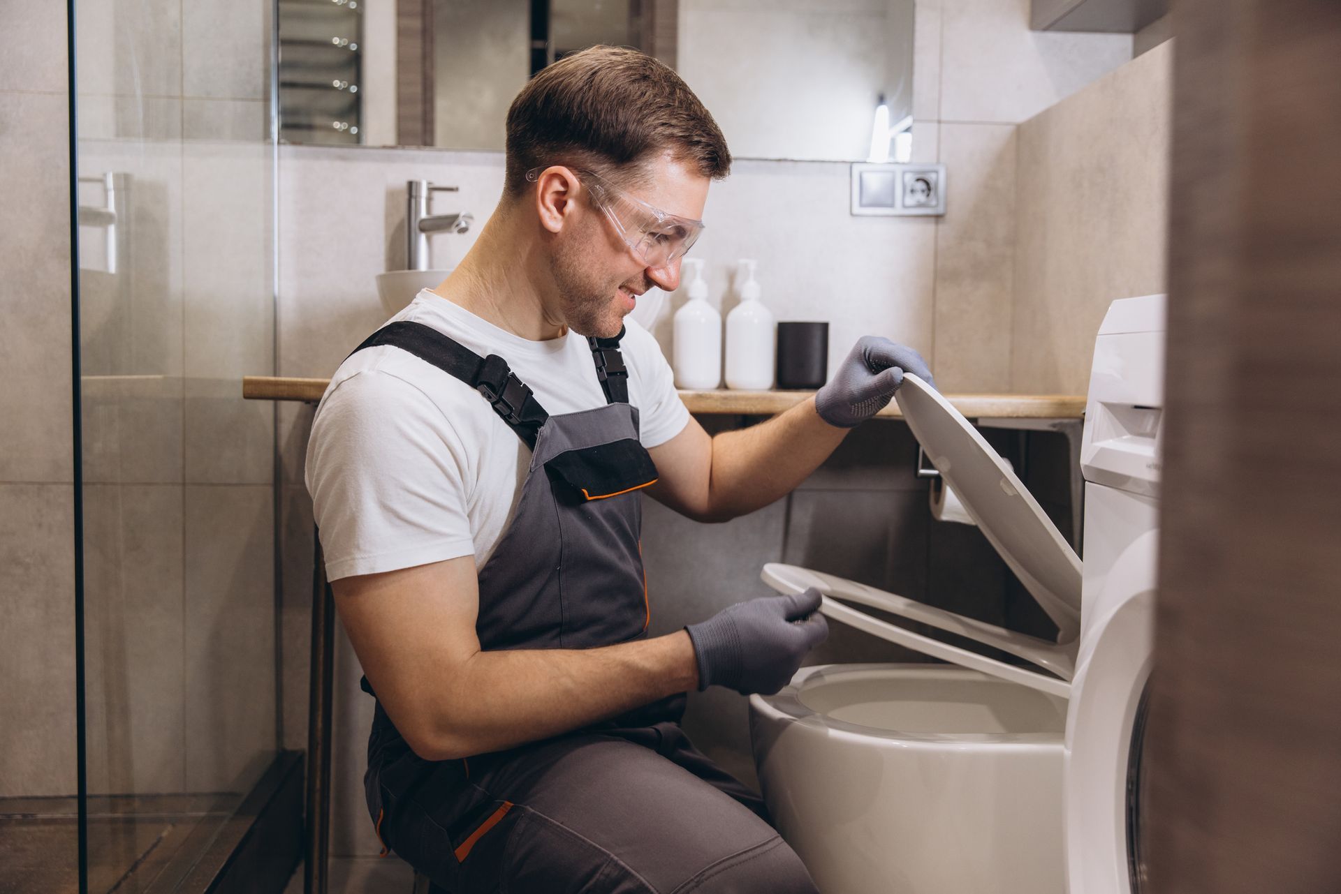 Man in work overalls and gloves examines a toilet in a bathroom.