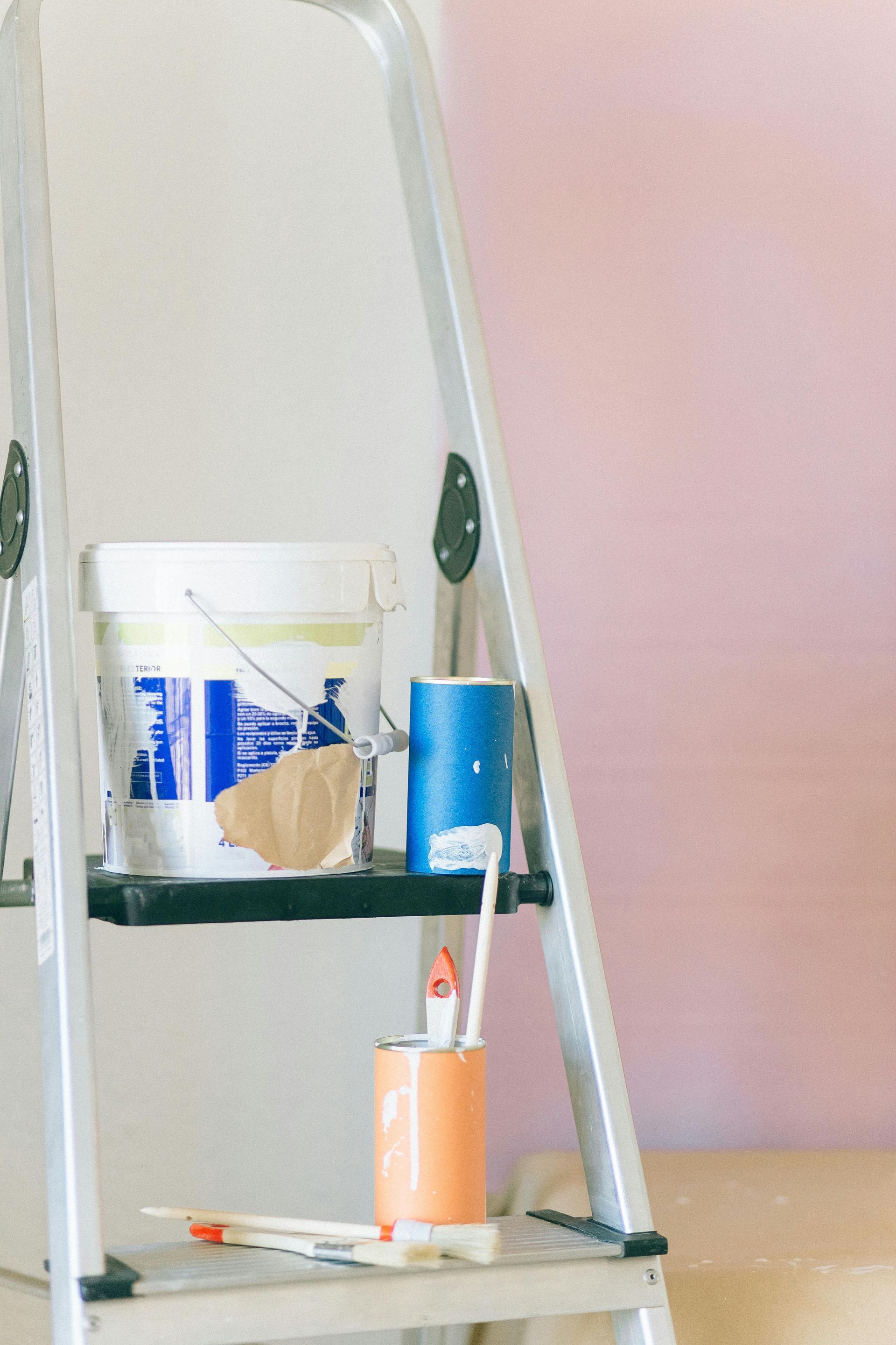 A ladder with a bucket of paint and brushes on it