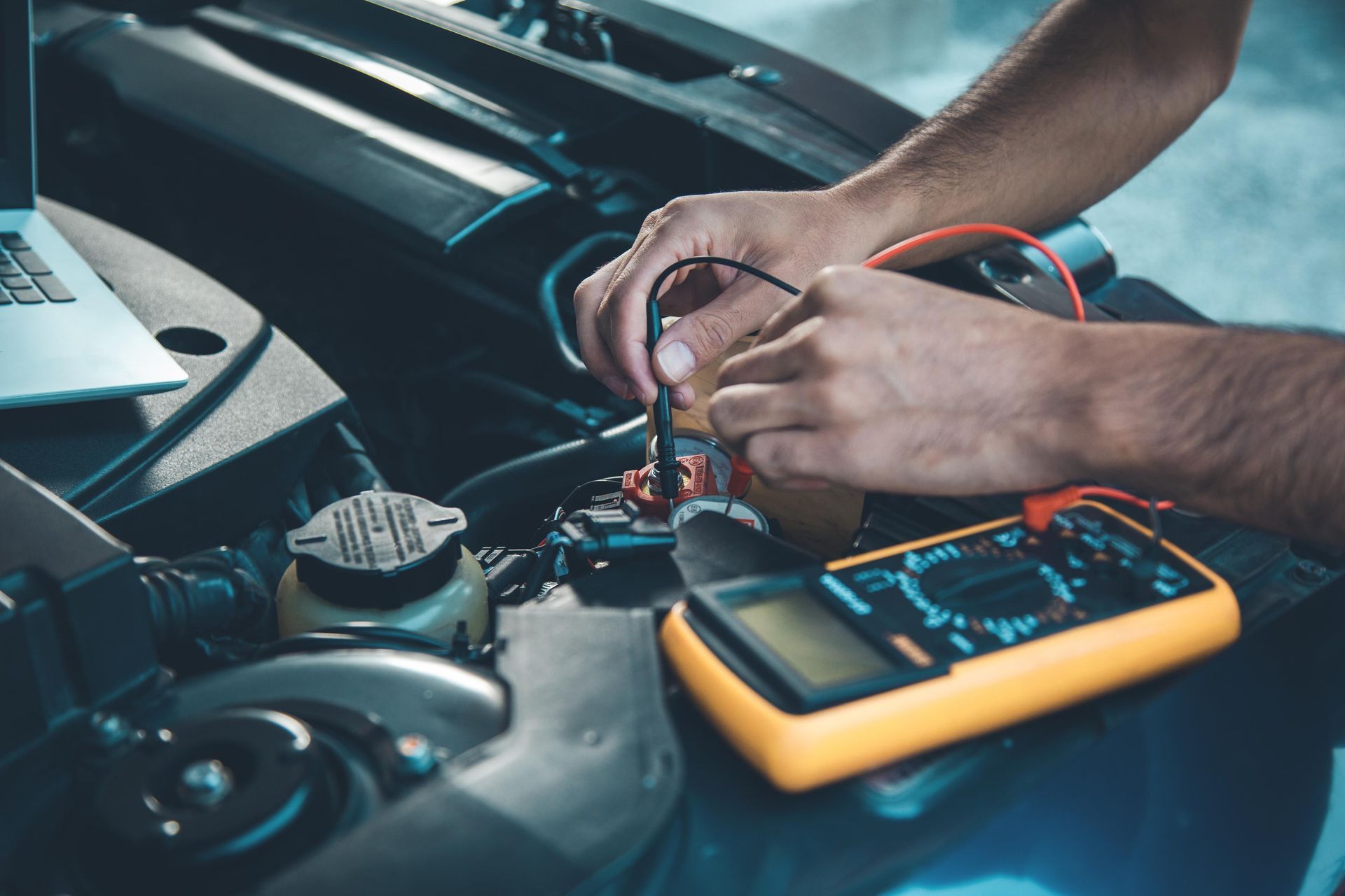 Hands testing a car's engine with a multimeter, laptop nearby. Automotive repair in progress.