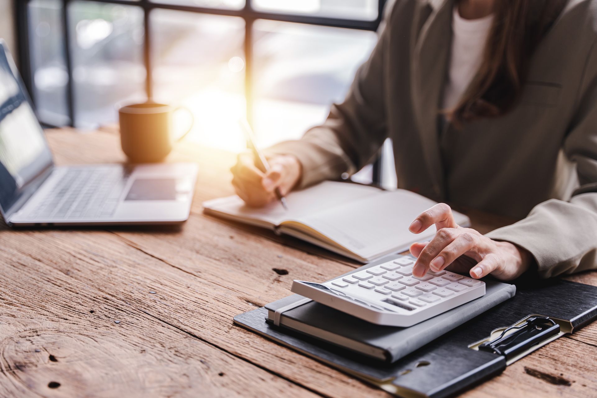 A woman is sitting at a wooden table using a calculator and writing in a notebook.