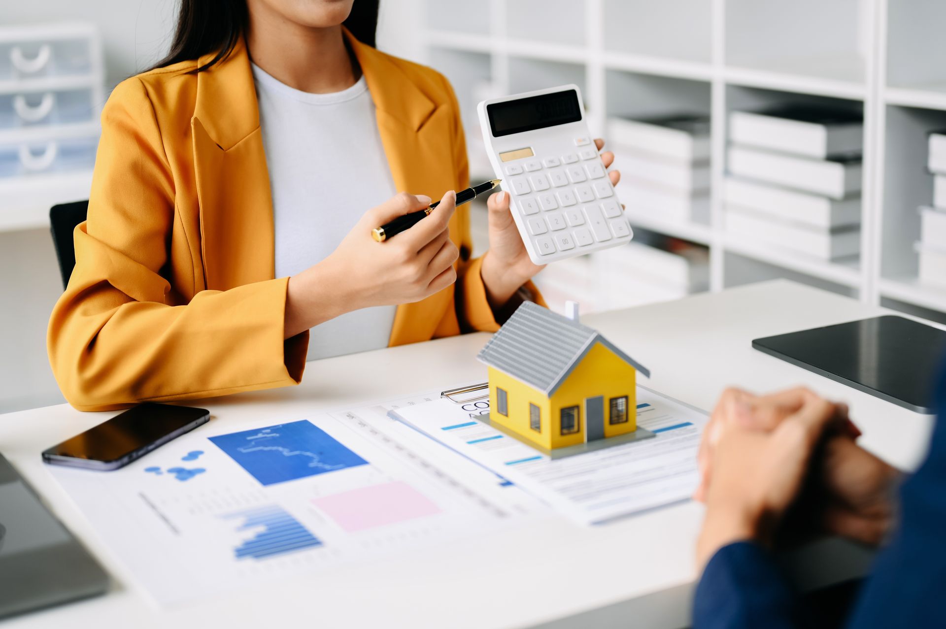 A woman is sitting at a table holding a calculator and a model house.