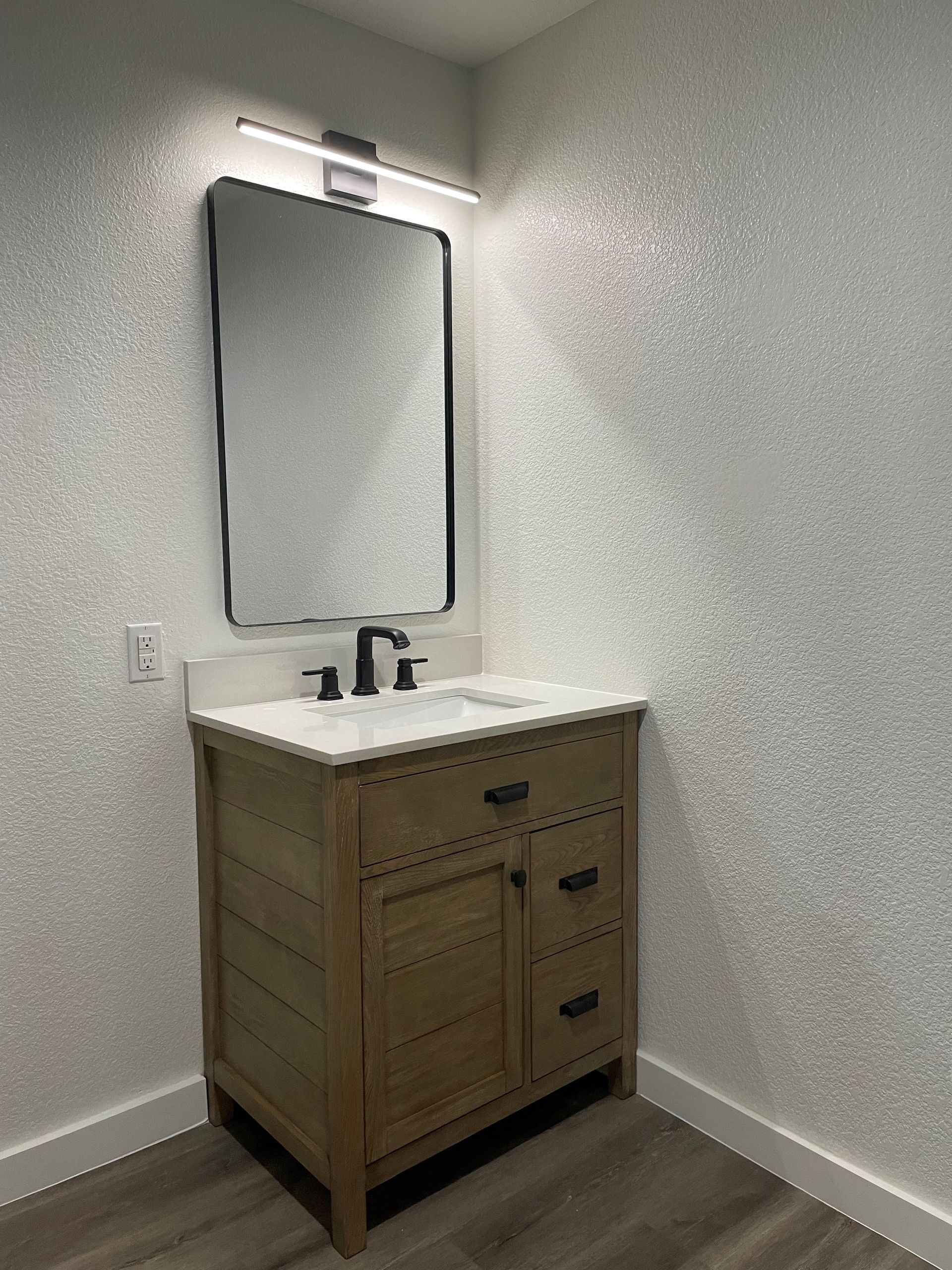 Bathroom vanity with wood cabinet, white countertop, black fixtures, and a mirror with light fixture.