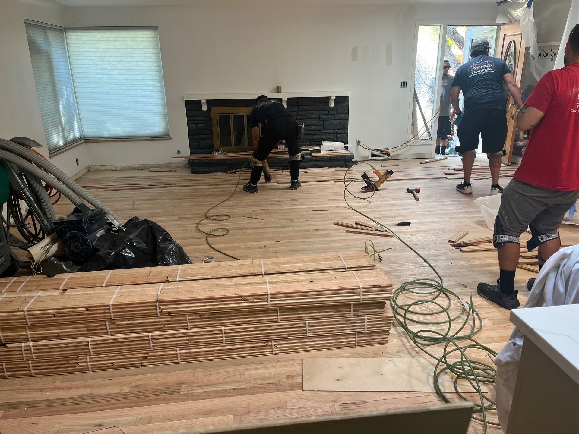 Men installing hardwood floors in a room with a fireplace. Boards stacked nearby.