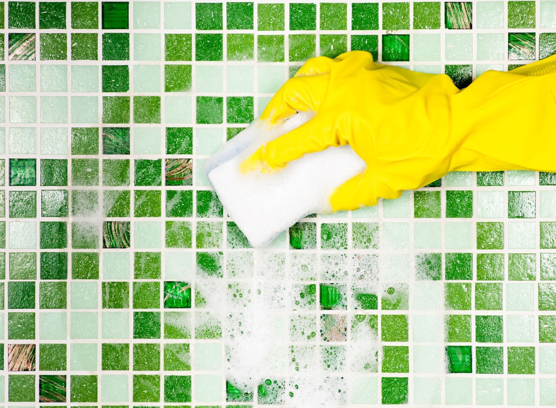A person wearing yellow gloves is cleaning a green tile wall with a sponge.
