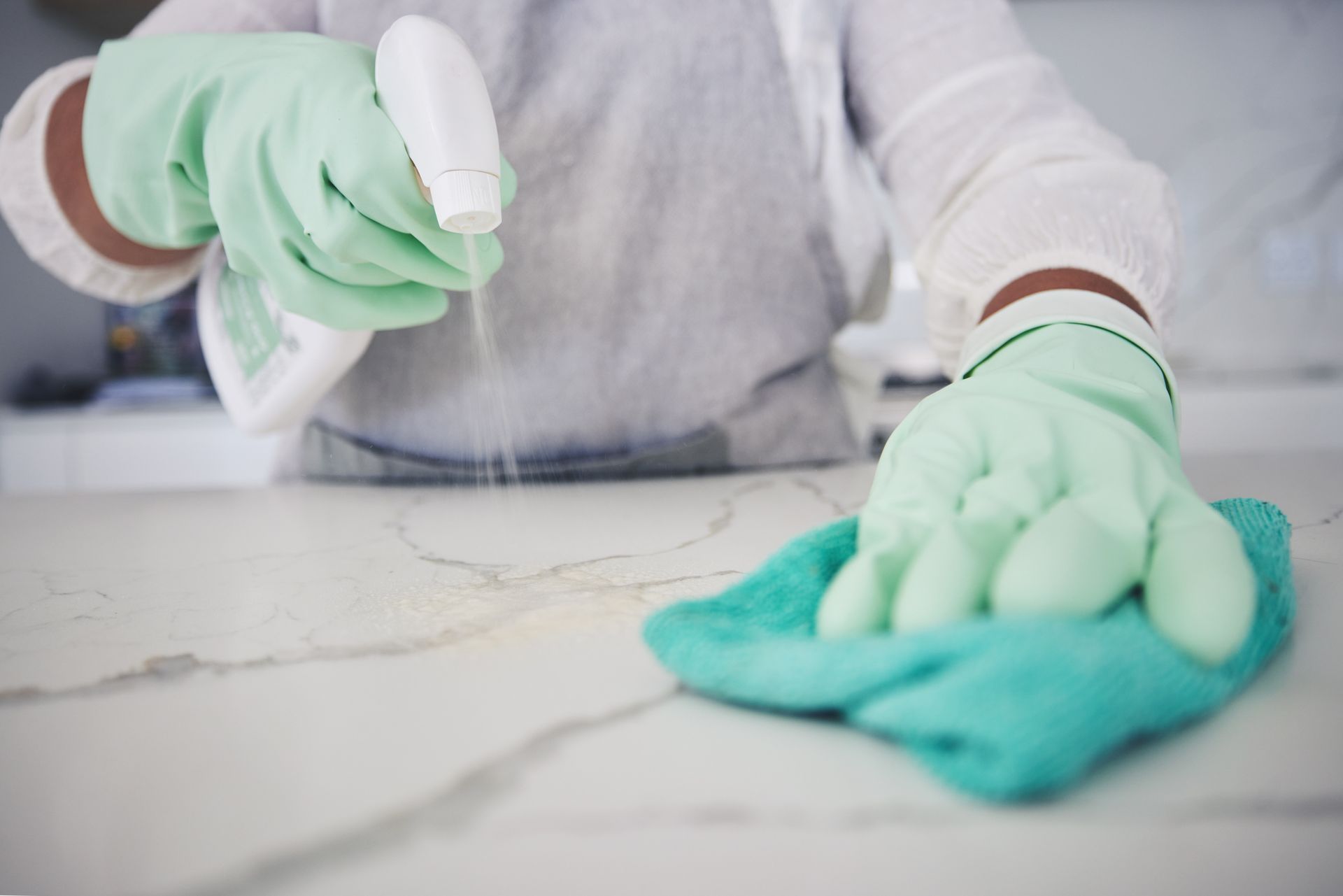 A person wearing green gloves is cleaning a counter with a cloth and spray bottle.