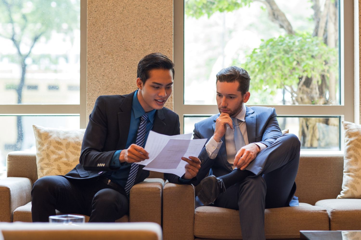 Two professionals in business suits sit on a sofa in a sunlit office, reviewing documents together.